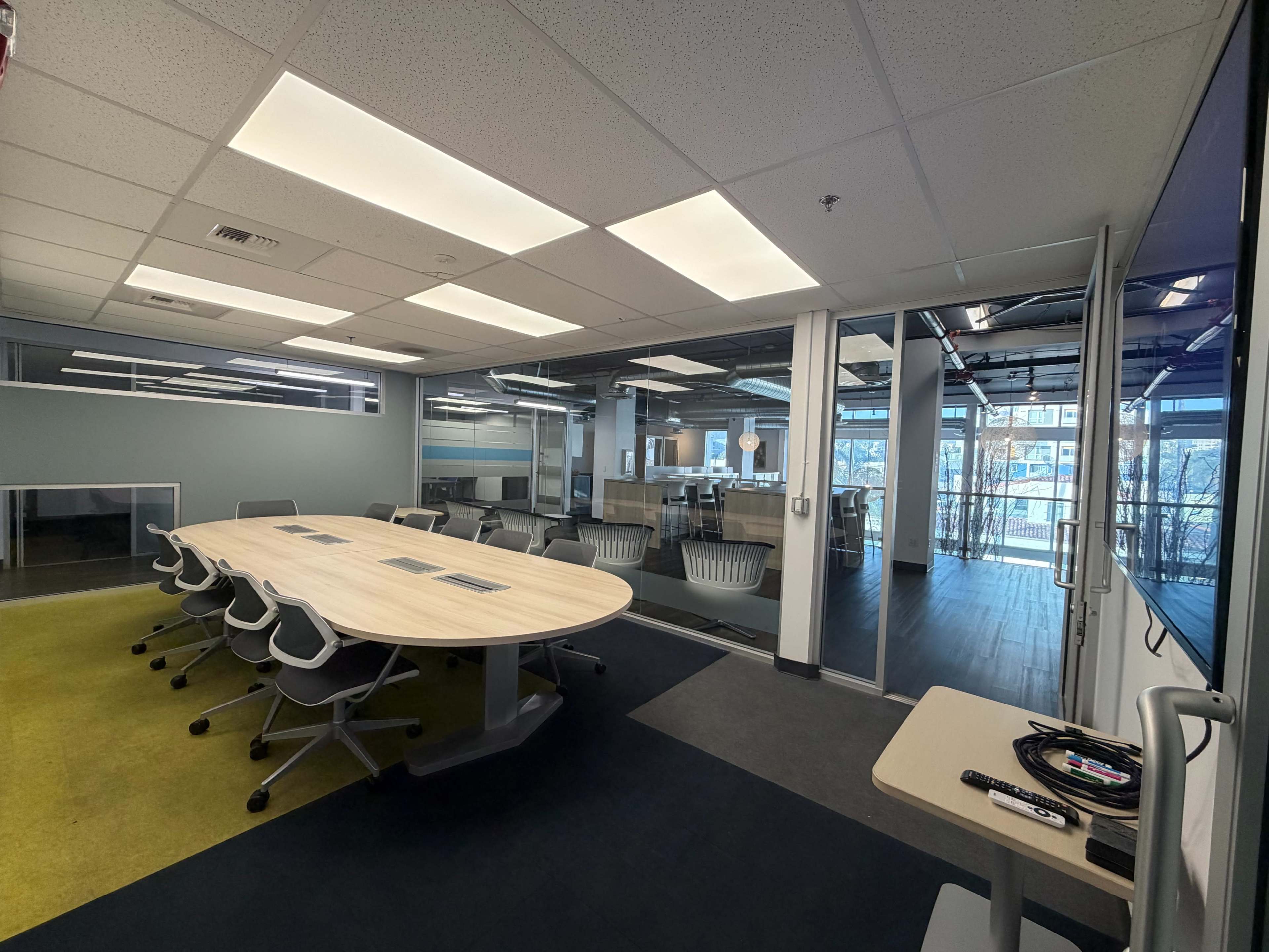 The image shows a modern conference room with a round table and large glass windows revealing another workspace.