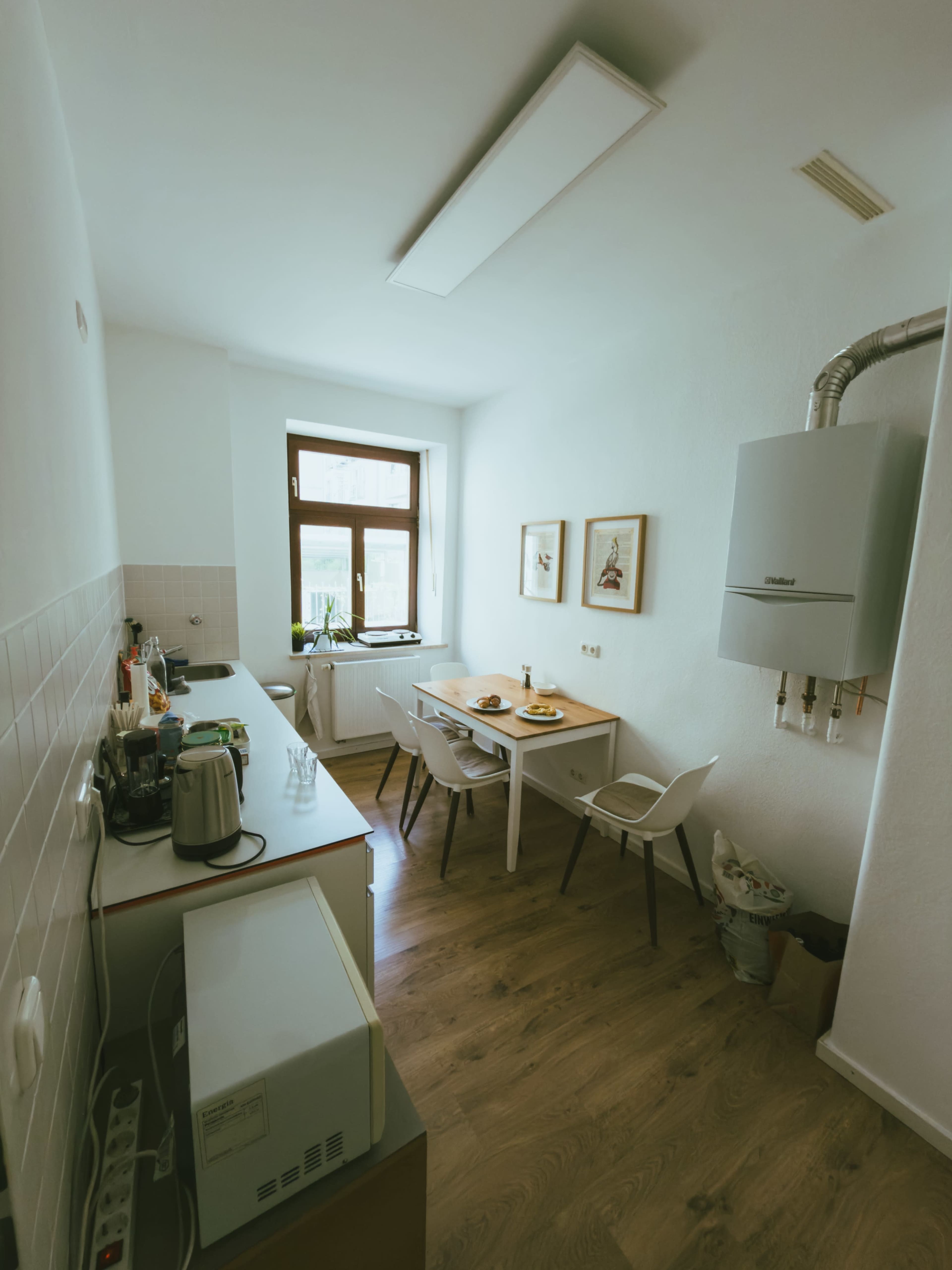 The image shows a compact kitchen with a table and chairs, a window, and appliances including a kettle and a microwave.