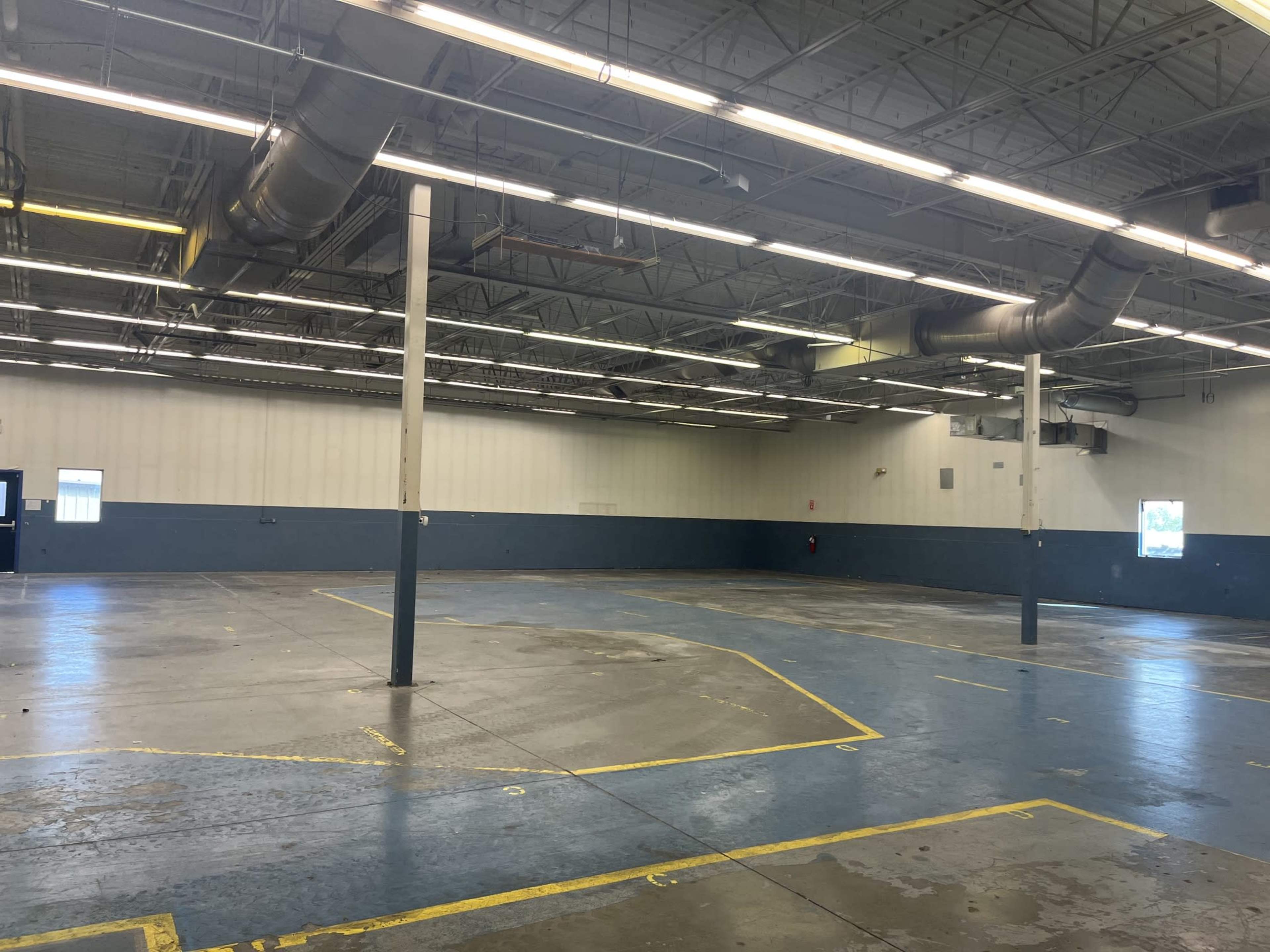 The image shows a large, empty industrial space with blue and gray walls, concrete flooring, and overhead ventilation ducts.