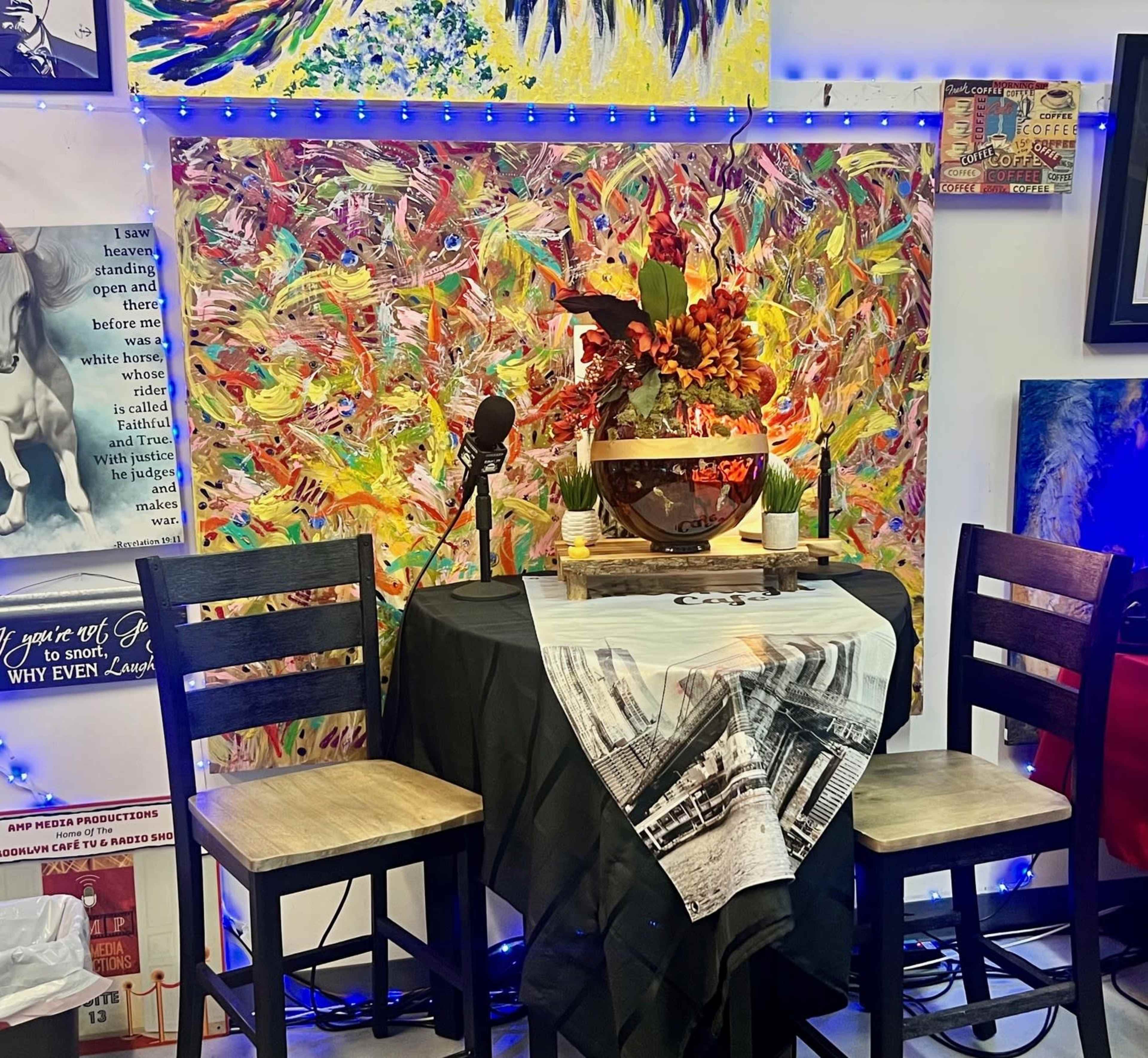A table set for two with a decorative bowl and flowers is positioned in front of a colorful abstract painting and accompanied by two wooden bar stools.