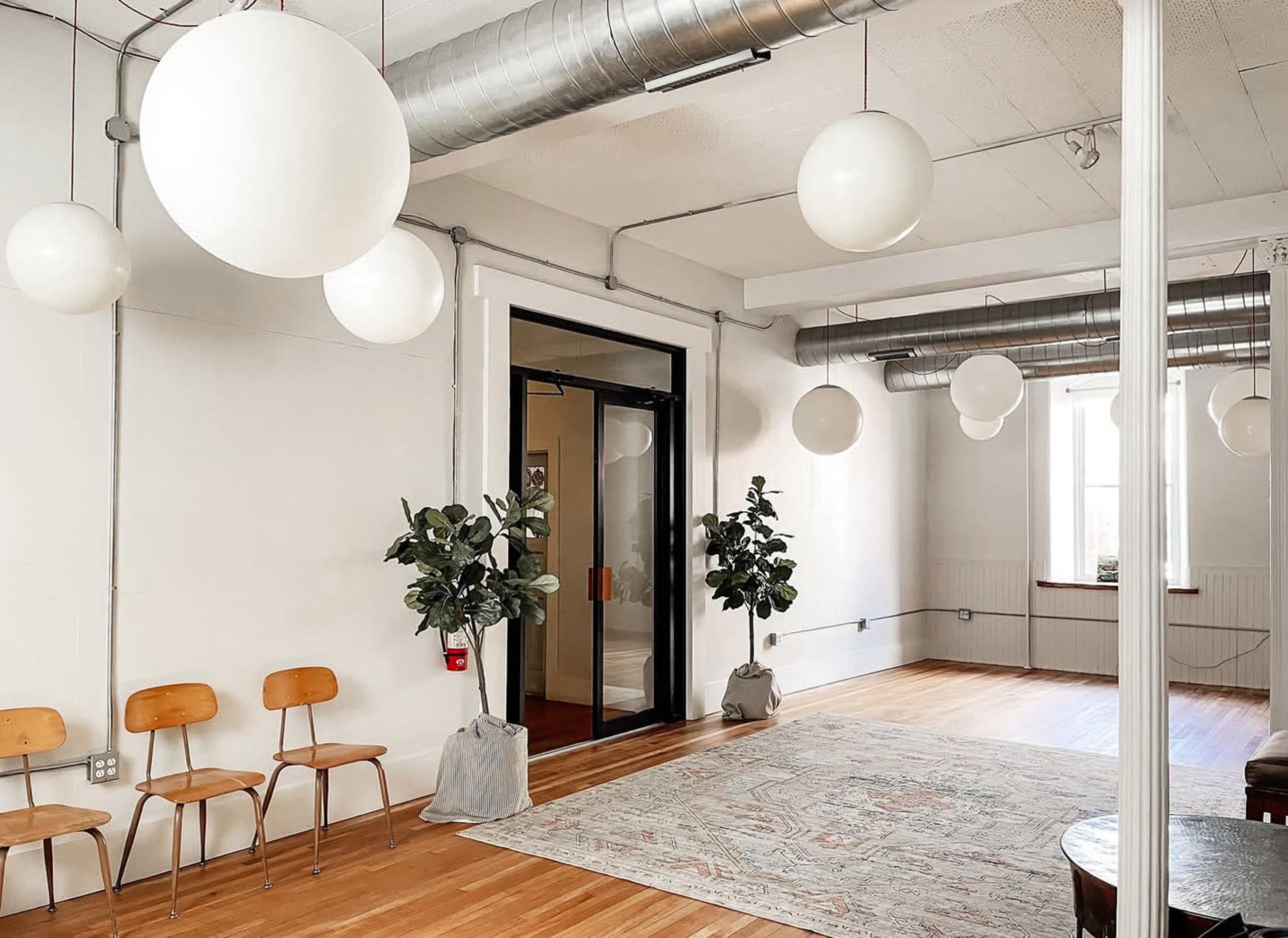 A bright, minimalist room features wooden floors, three wooden chairs, two potted plants, and several large white pendant lights hanging from the ceiling.