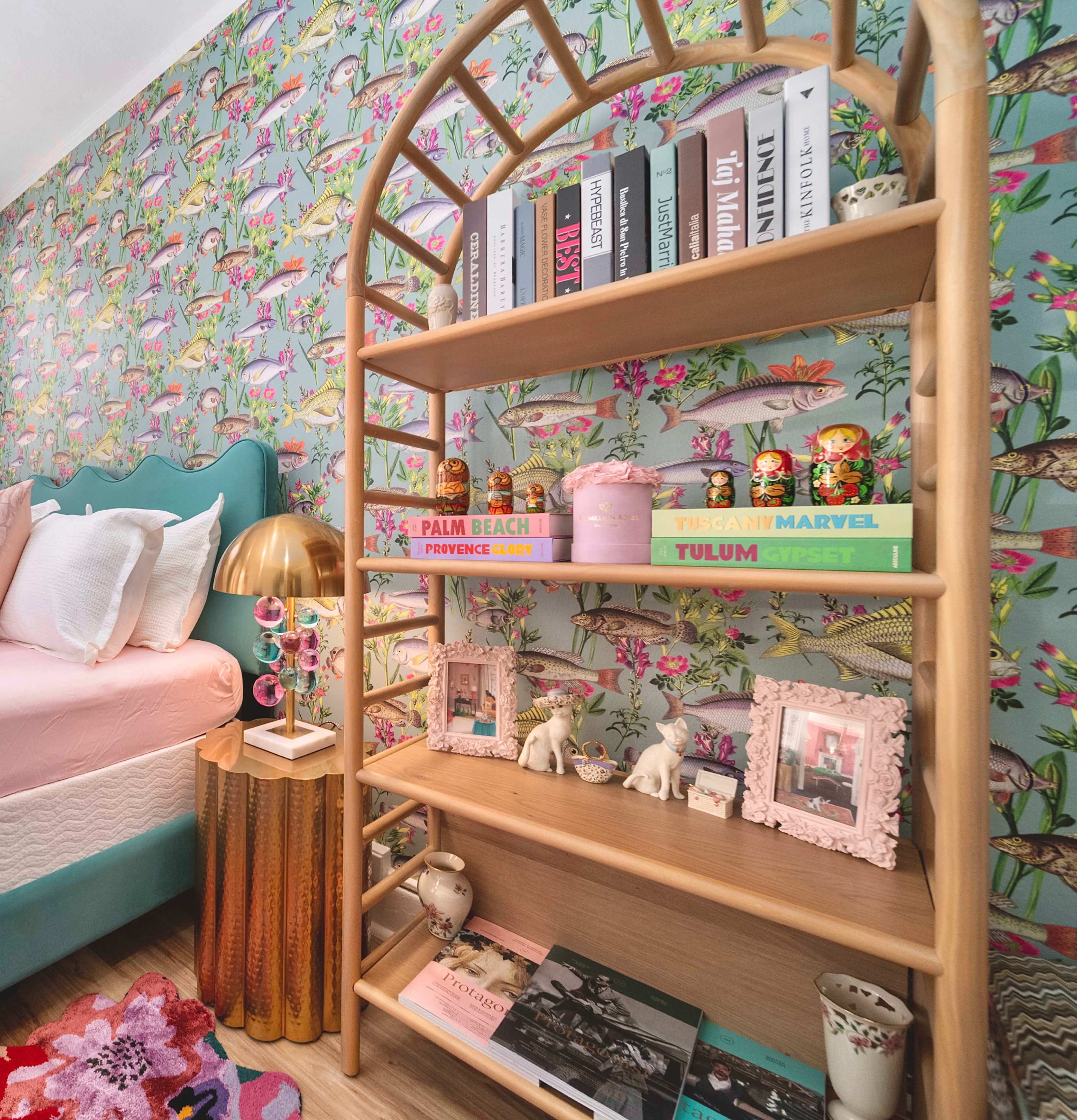 A colorful bedroom with a wooden bookshelf displaying various decorative items, books, and framed photos against a floral wallpaper backdrop.