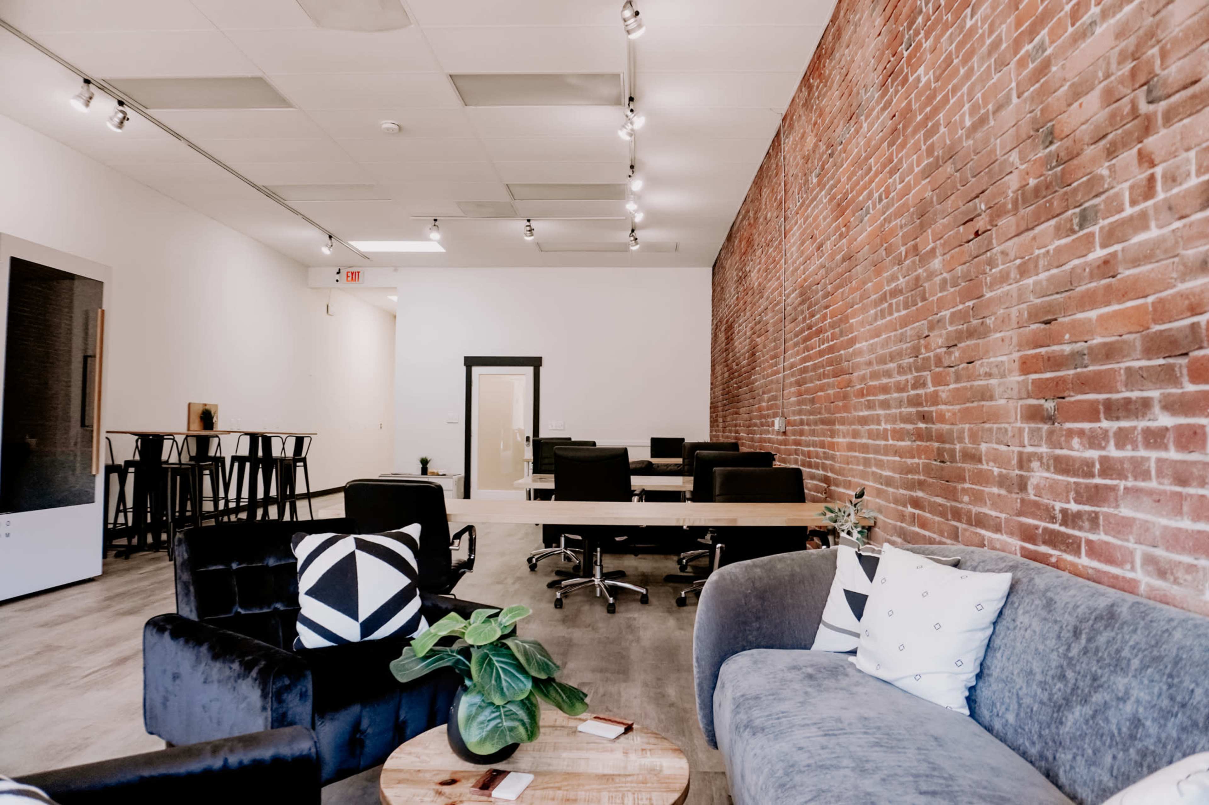 A modern office space featuring a combination of a brick wall, workstations with chairs, and a lounge area with couches and plants.