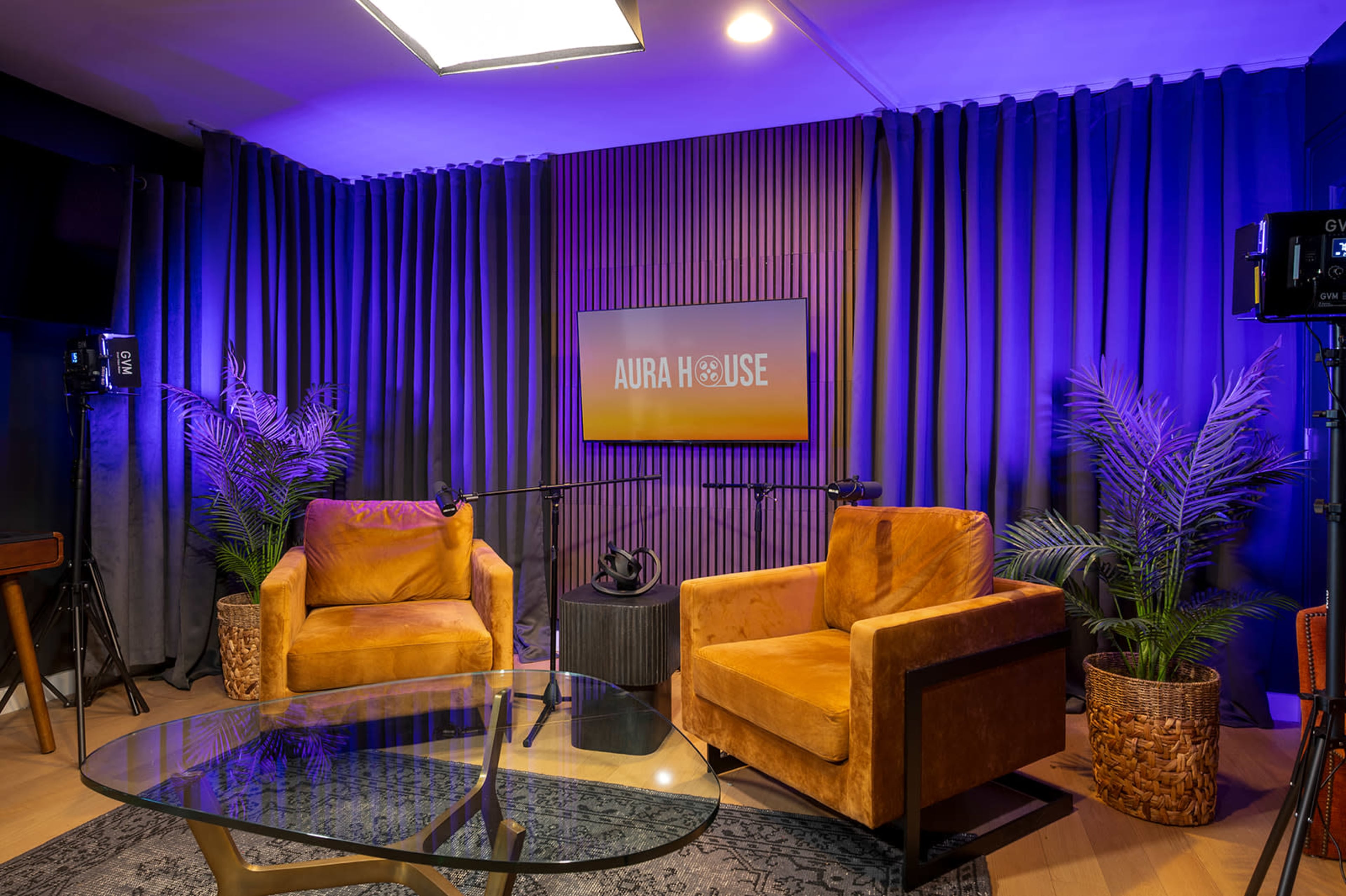 The image shows a styled interview or podcast setup featuring two brown chairs, a round glass table, and a backdrop with the text "AURA HOUSE," along with accent lighting and potted plants.
