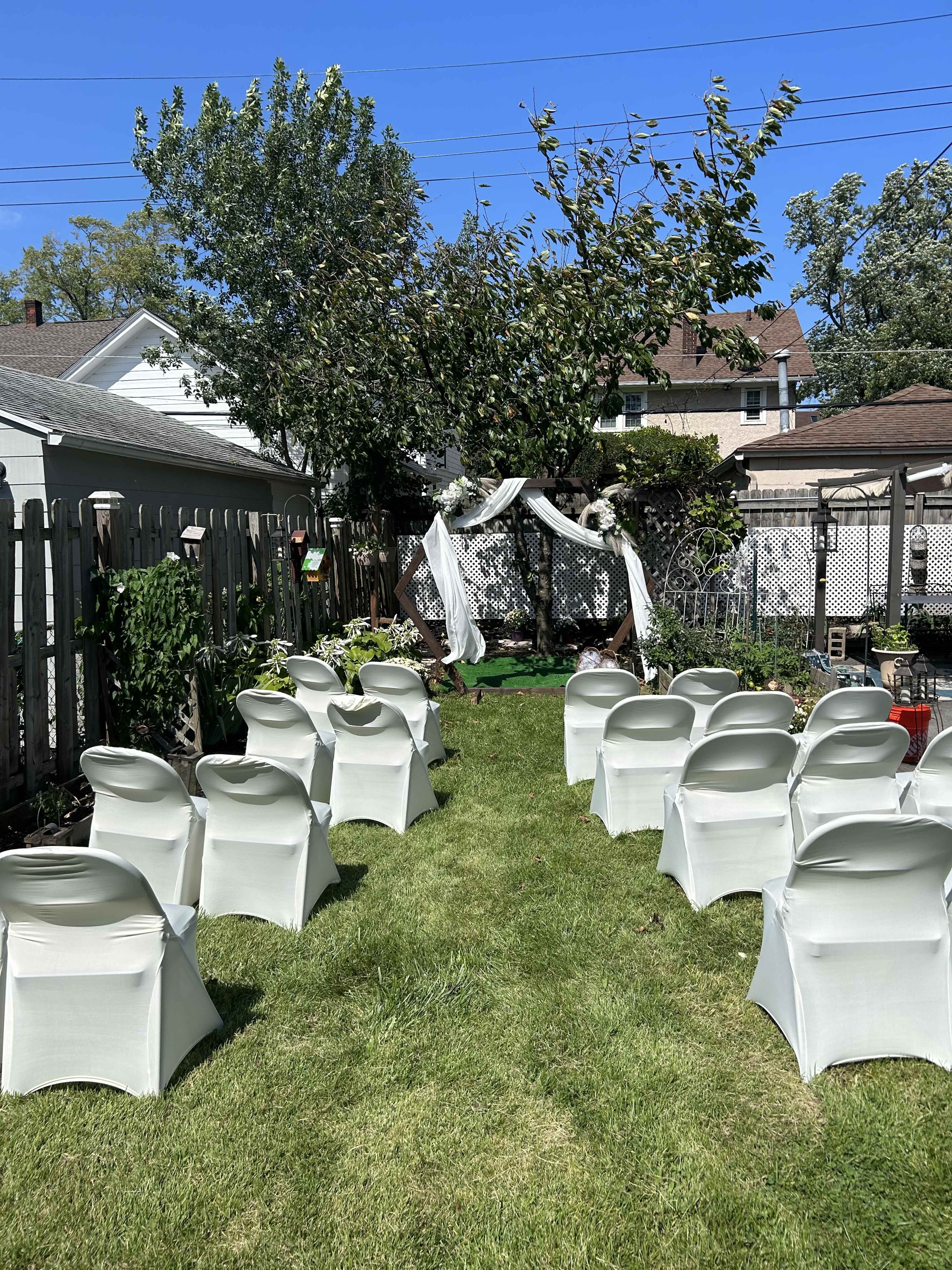 A garden is set up for a ceremony with white chairs lined up on a grassy area facing a decorated arch under a tree.