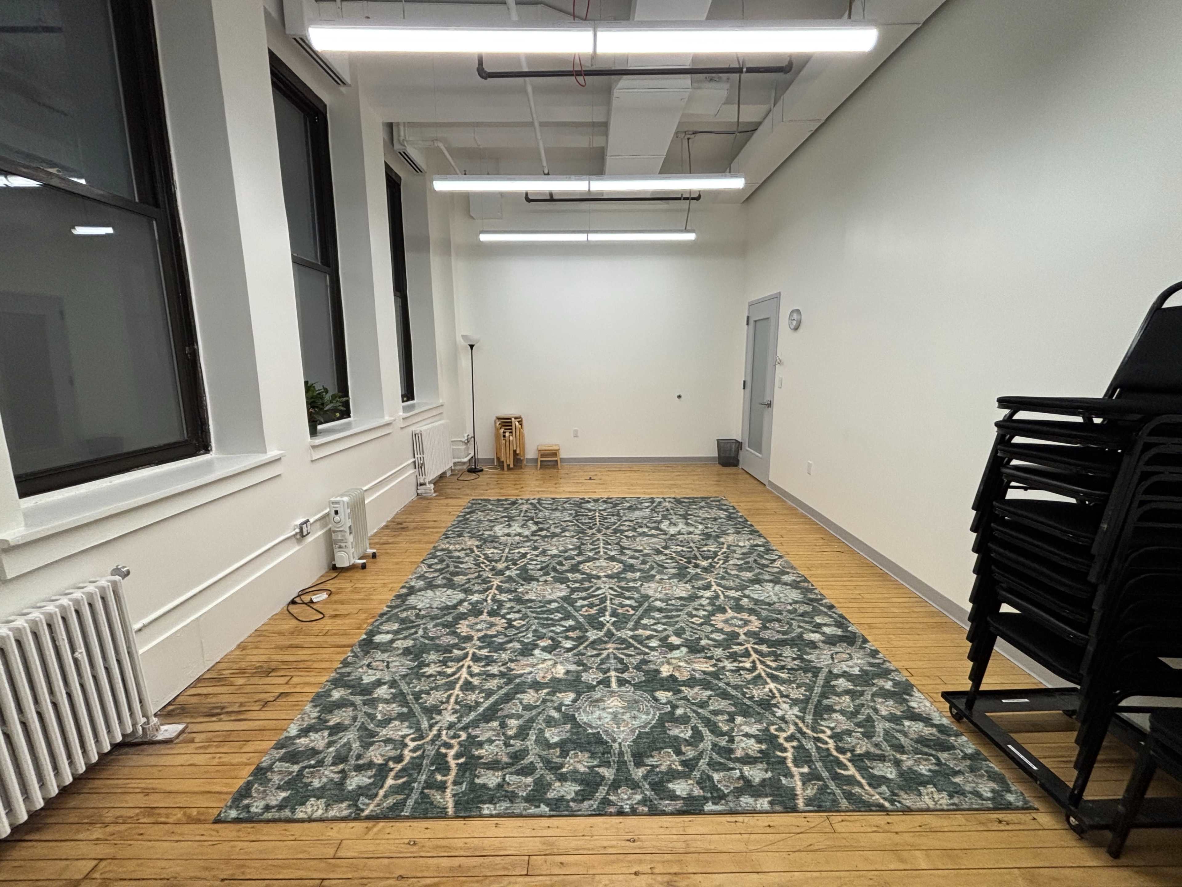 The image shows an empty room with wooden flooring, a large area rug in the center, and a stack of chairs against one wall.