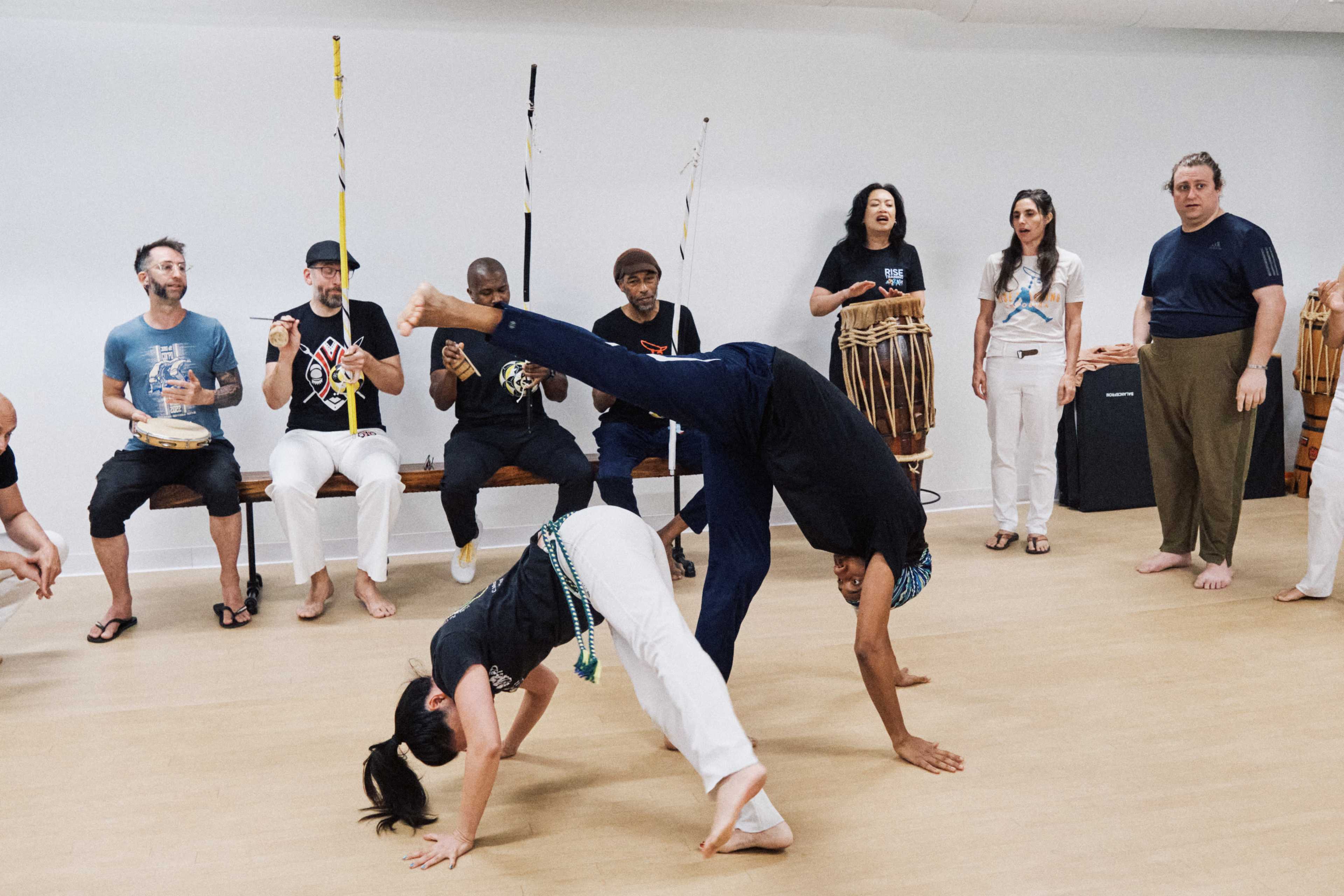 A group of people is participating in a capoeira class, with some individuals performing acrobatic movements while others play instruments and sing.