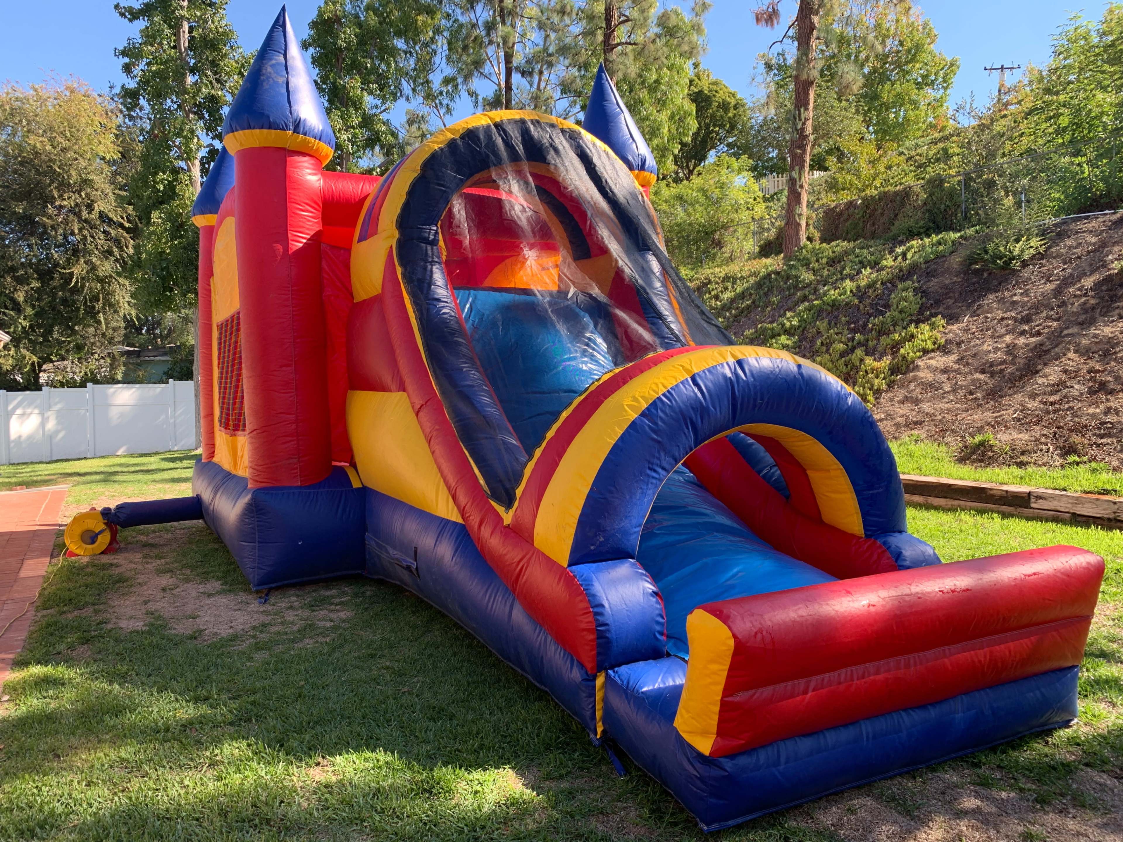 A colorful inflatable bounce house with a slide is set up on a grassy area surrounded by trees.