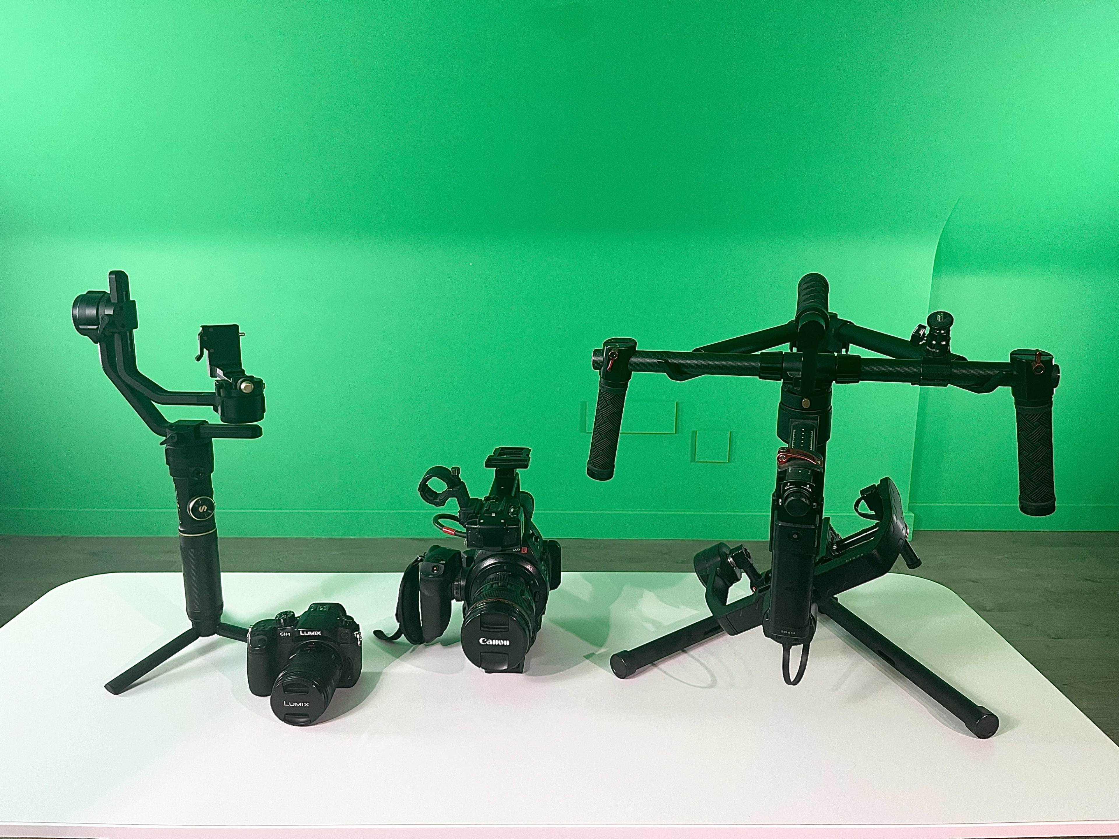 Three camera stabilizers and two Canon cameras arranged on a white table with a green background.