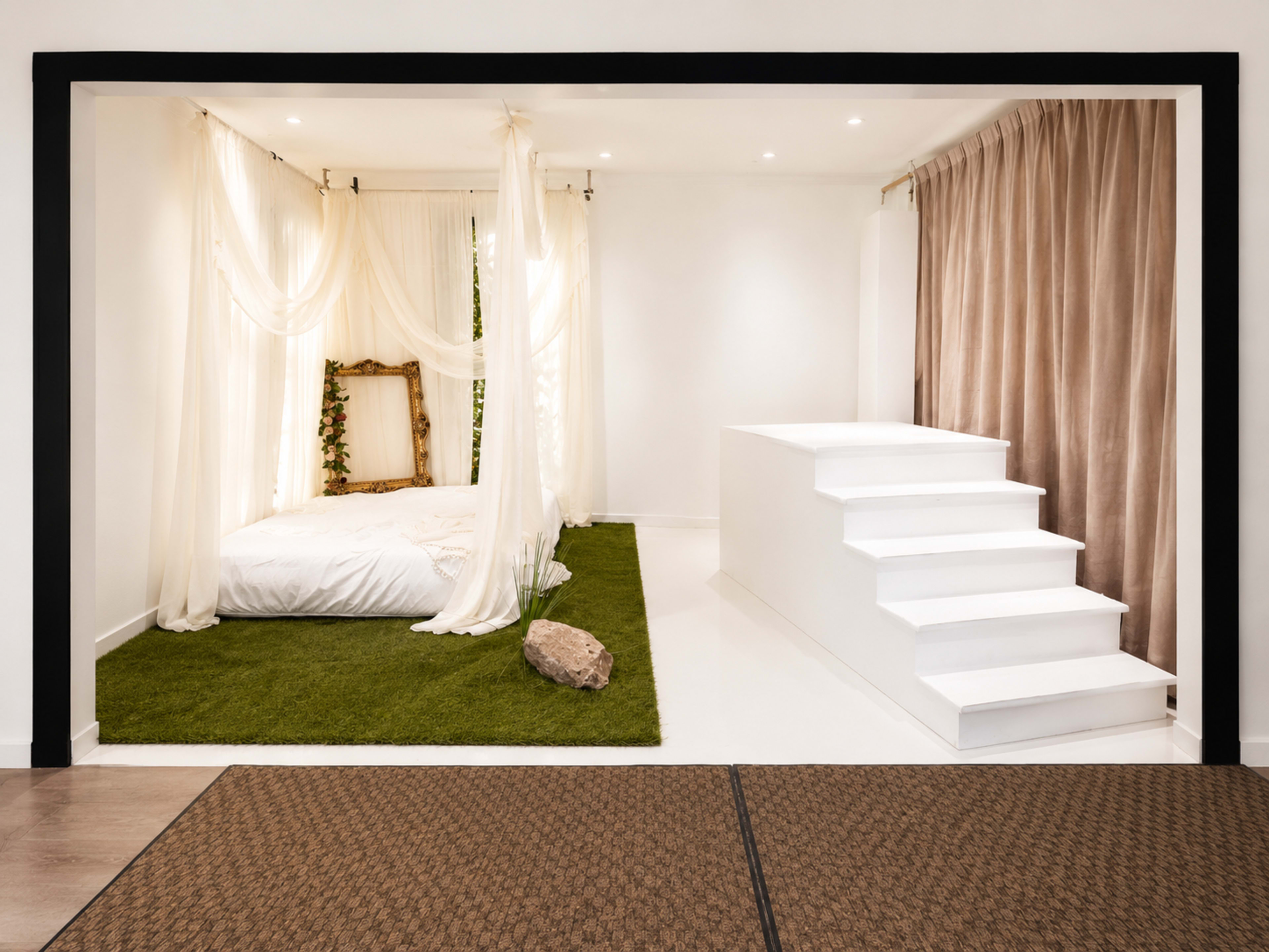 The image shows a modern bedroom featuring a bed with draped fabric, a large mirror, artificial grass, and a set of white stairs leading to a raised platform.