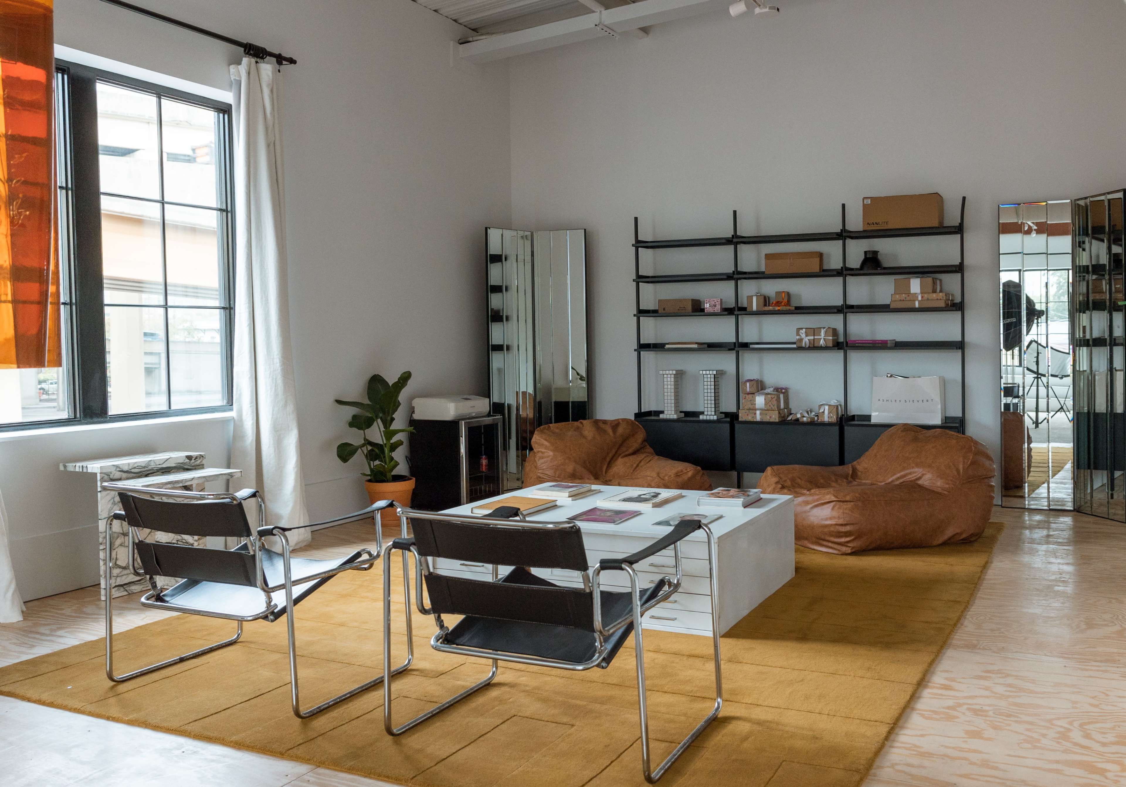 The image shows a modern room with a seating area featuring two chairs, a large table, and two bean bag chairs, alongside shelves filled with various items and a plant in the corner.