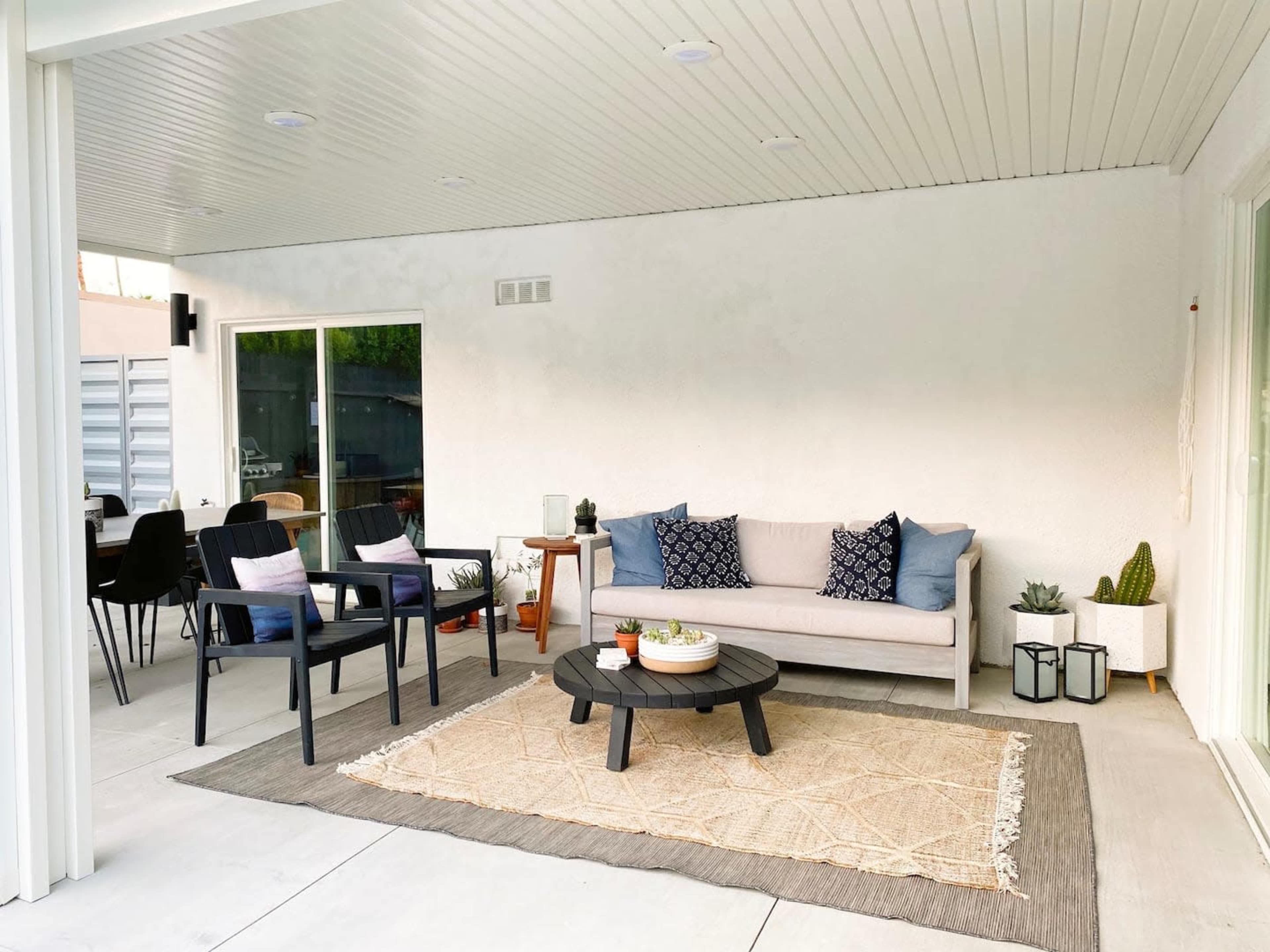 A covered outdoor seating area featuring a couch, two chairs, a round coffee table, and decorative plants on a light-colored rug.