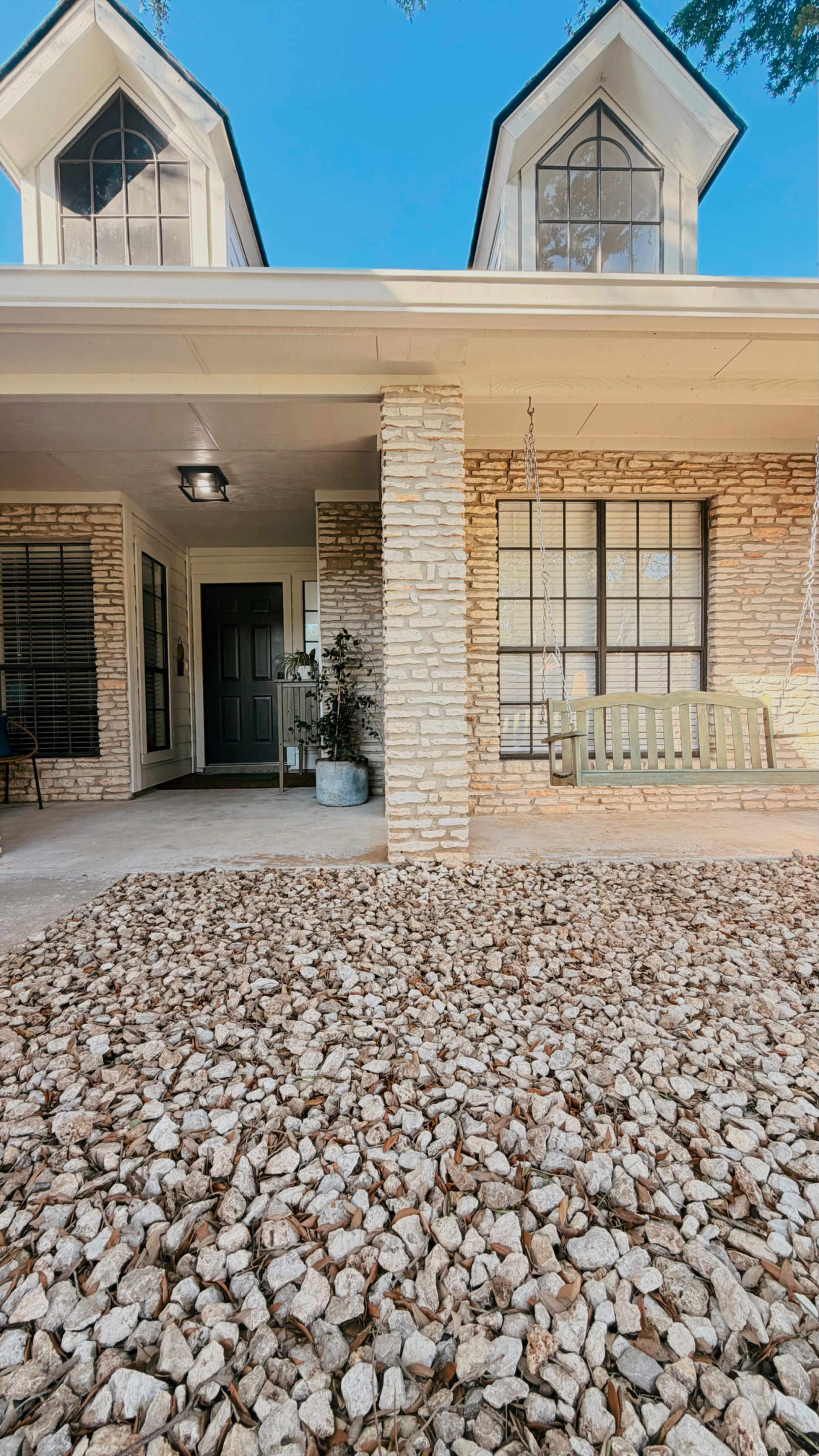 The image shows a house exterior with a front porch featuring a swing, a stone column, and a landscaped area covered in small rocks.