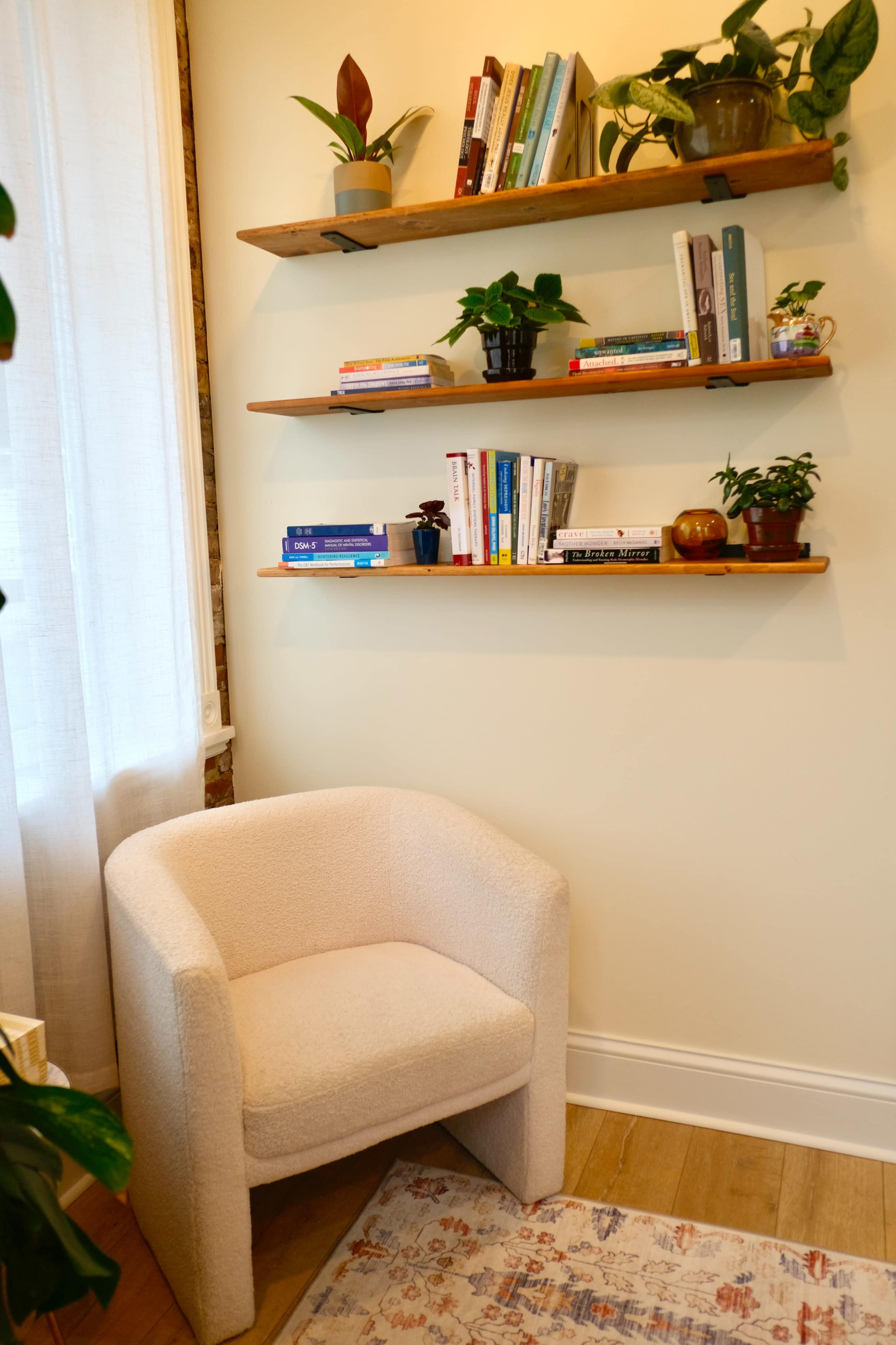 The image shows a cozy corner of a room featuring a soft, upholstered chair next to three wooden shelves filled with books and potted plants.