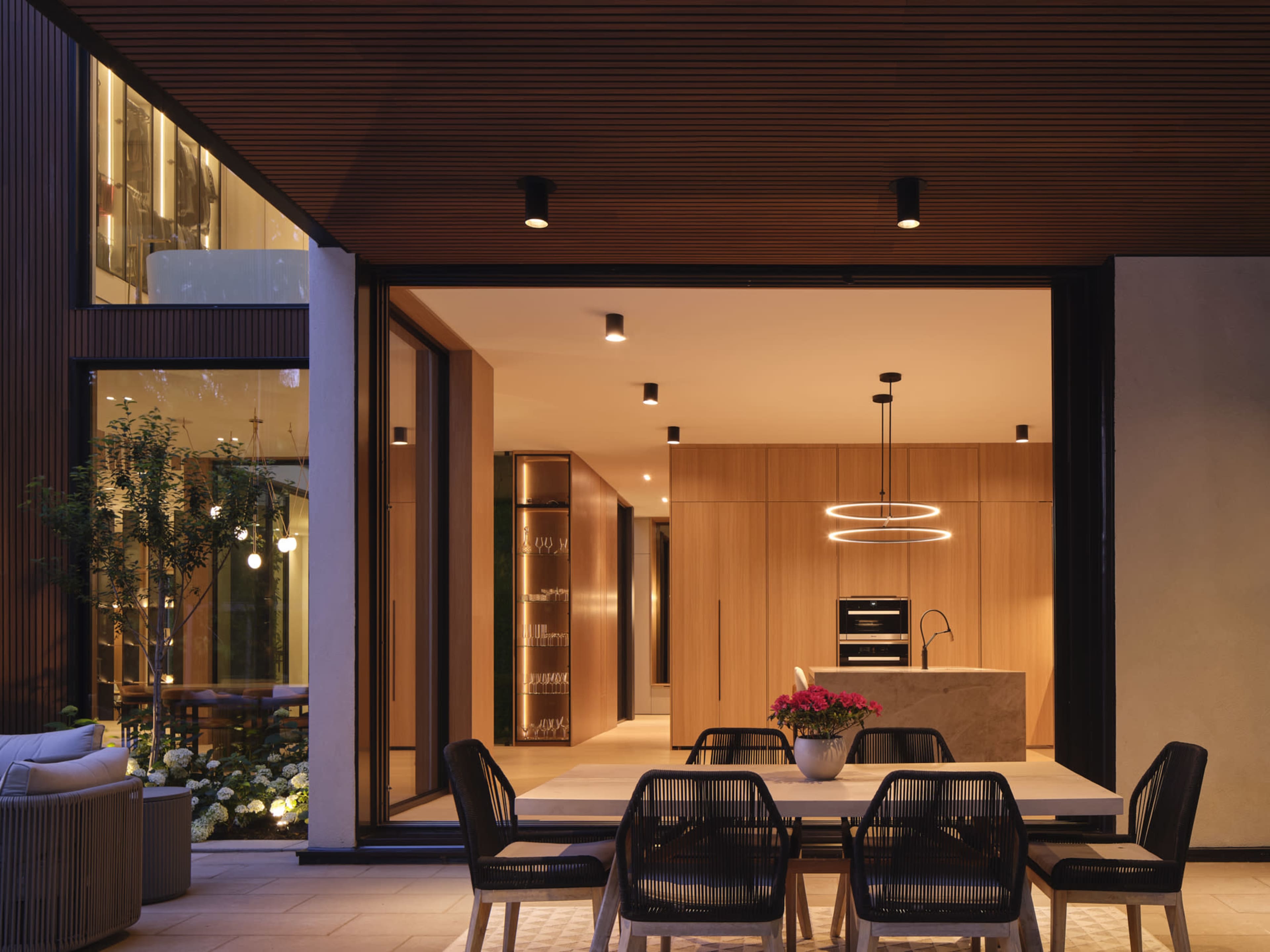 The image shows a modern patio area with a dining table and four chairs, overlooking a contemporary kitchen featuring wooden cabinetry and an island with a circular pendant light.