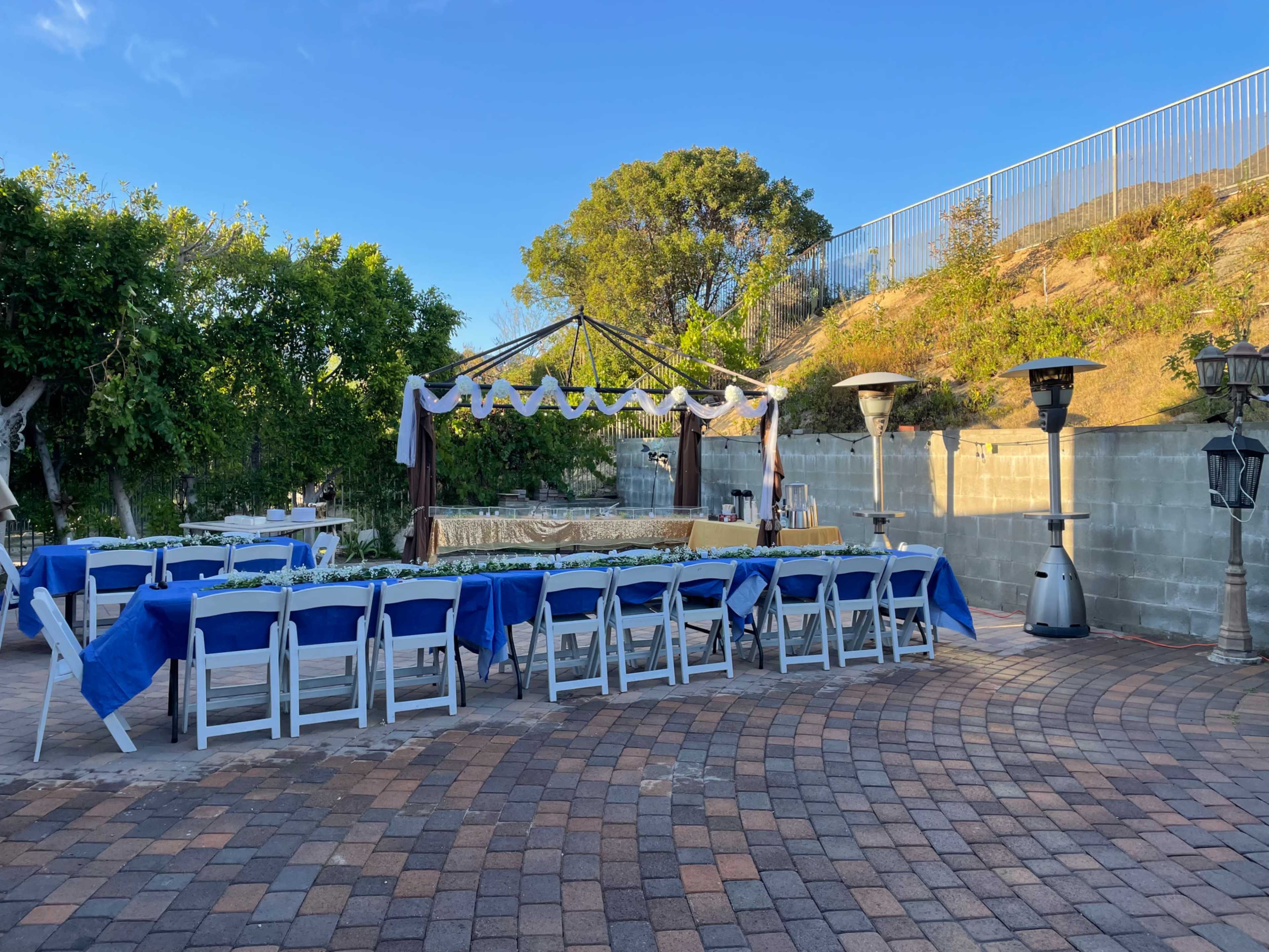 A set-up for an outdoor event features rows of blue-covered tables and a decorated canopy in a garden area.