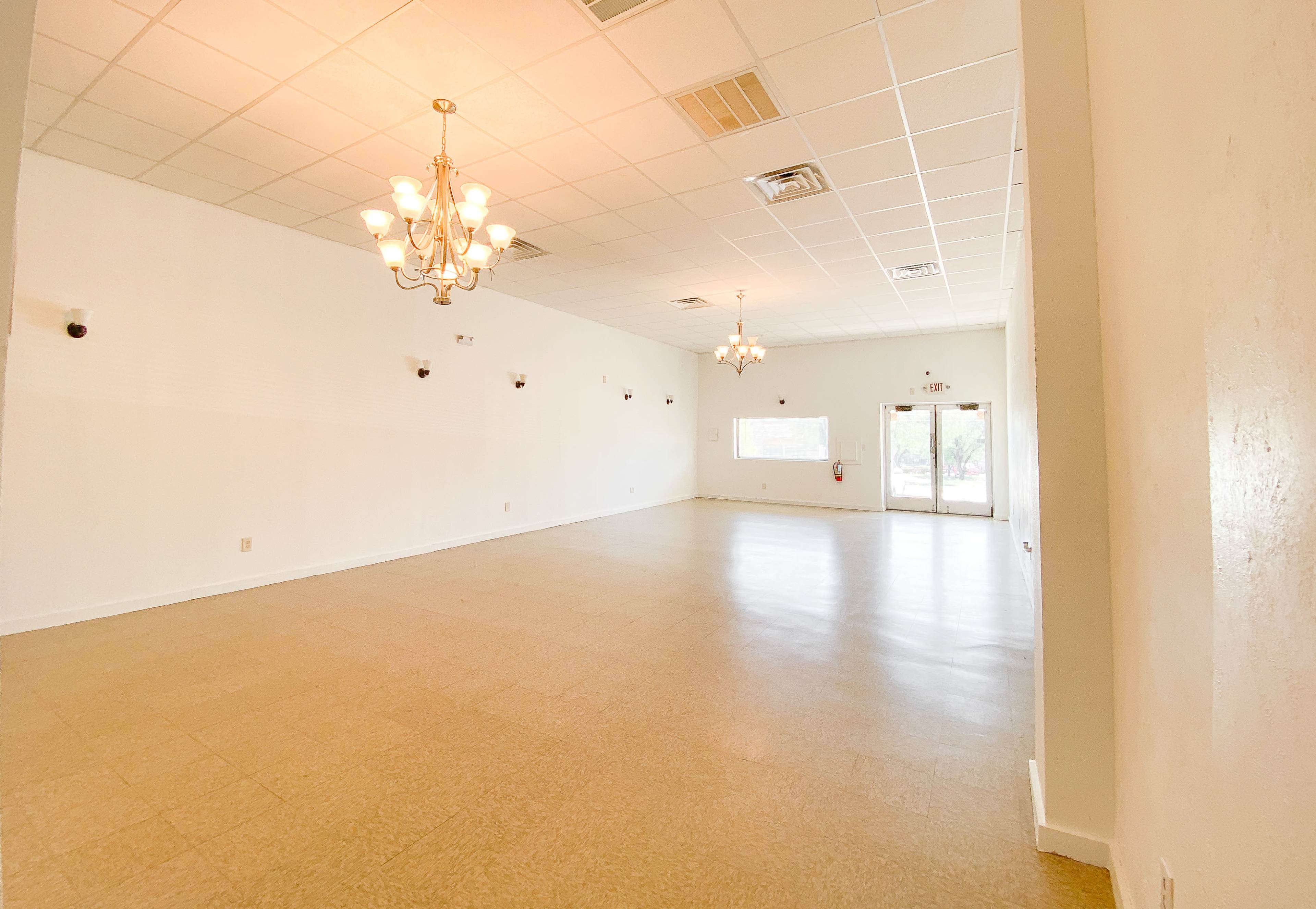 The image shows a spacious, empty room featuring a light-colored floor and ceiling lights, with large windows at one end and a door leading outside.