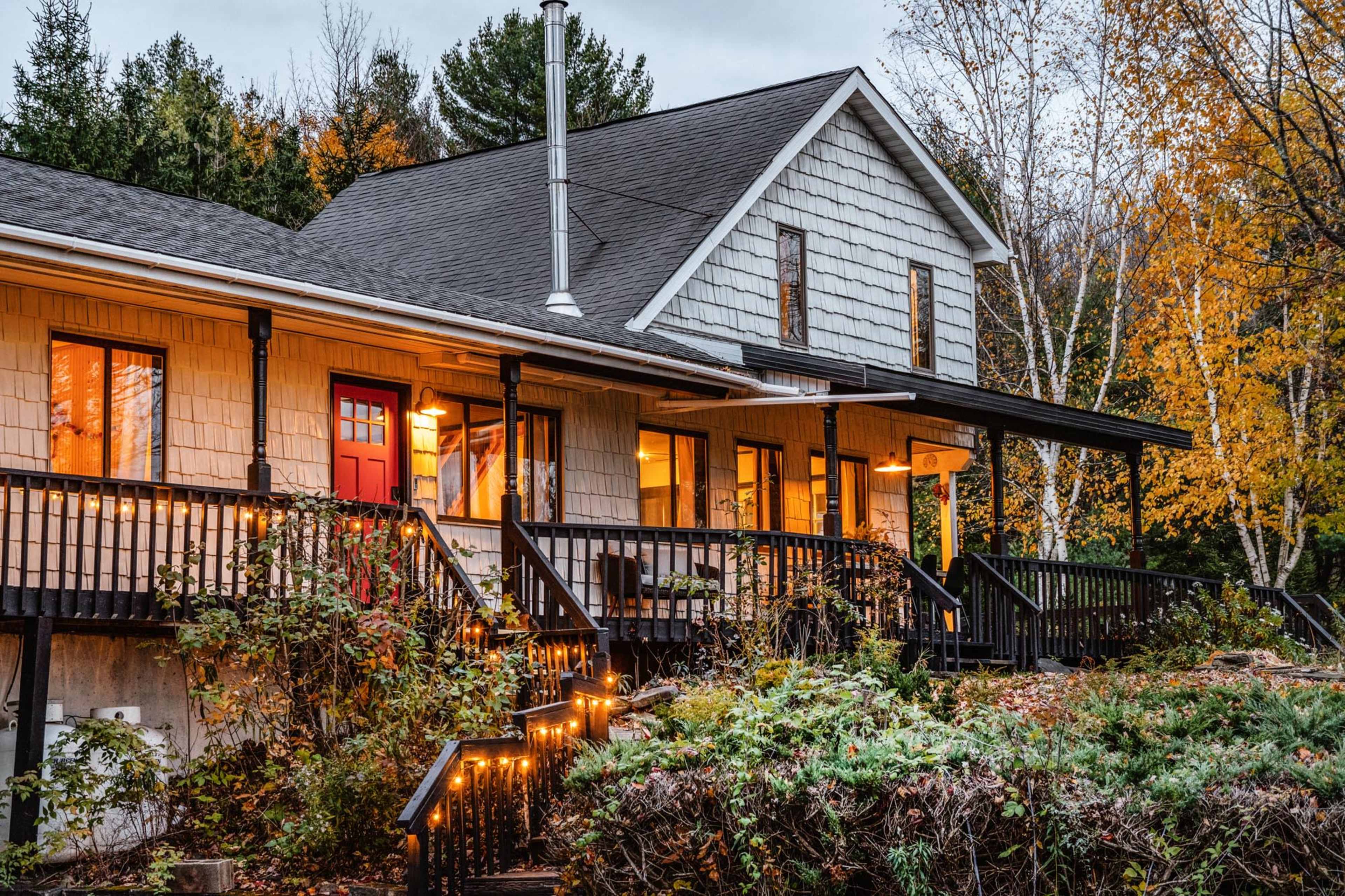 A house with a shingled facade and red door is surrounded by autumn foliage and features a front porch with lights and wooden railings.