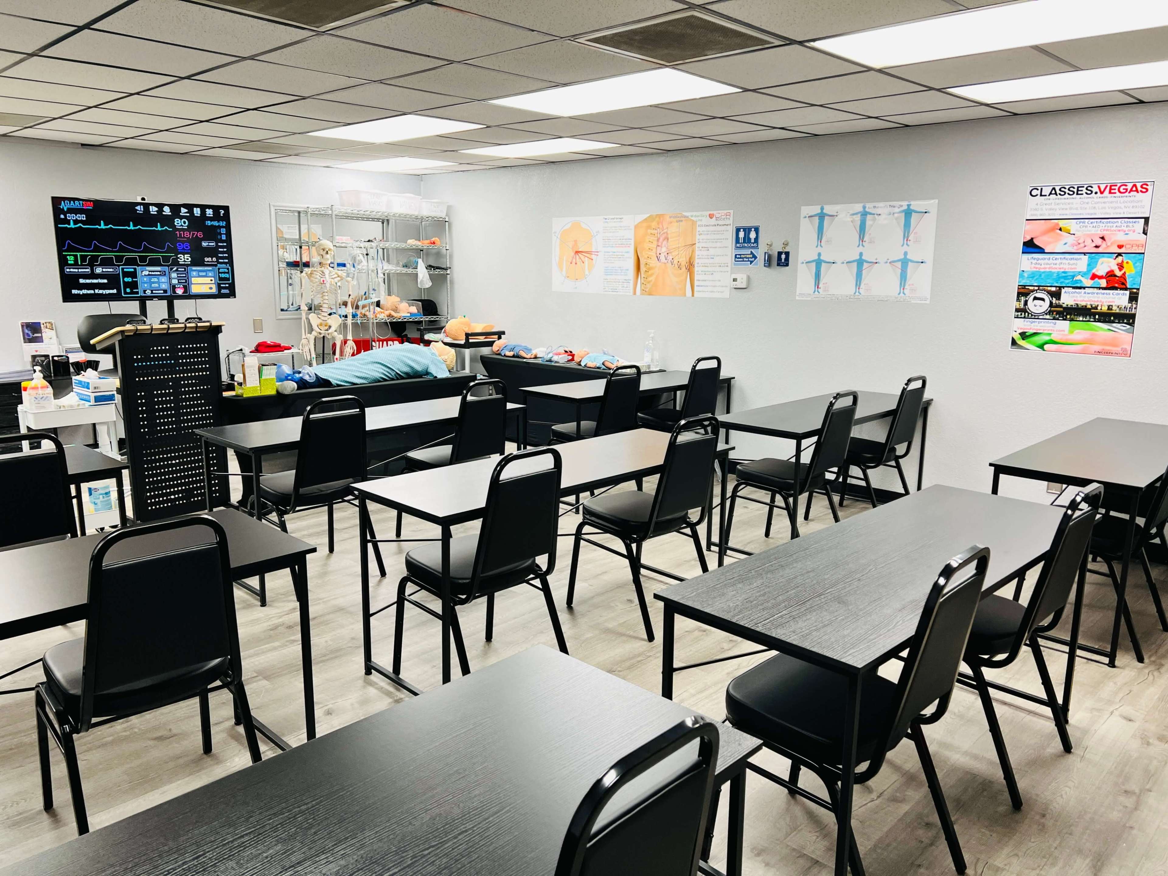 The image shows a classroom setup with multiple black tables and chairs, featuring medical equipment and instructional posters on the walls.