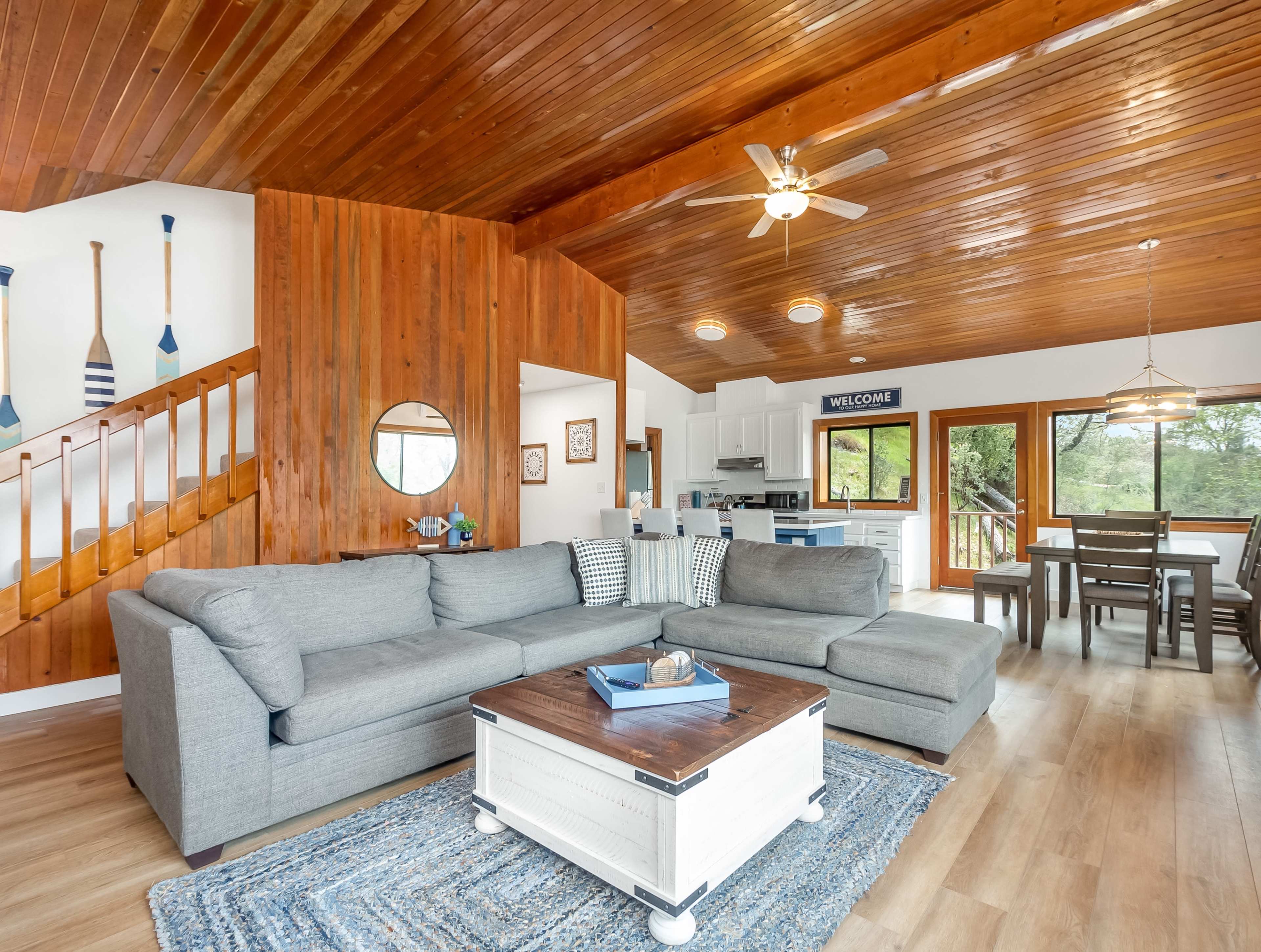 A cozy living room with a gray sectional sofa, a white coffee table, wooden ceiling details, and large windows overlooking a wooded area.