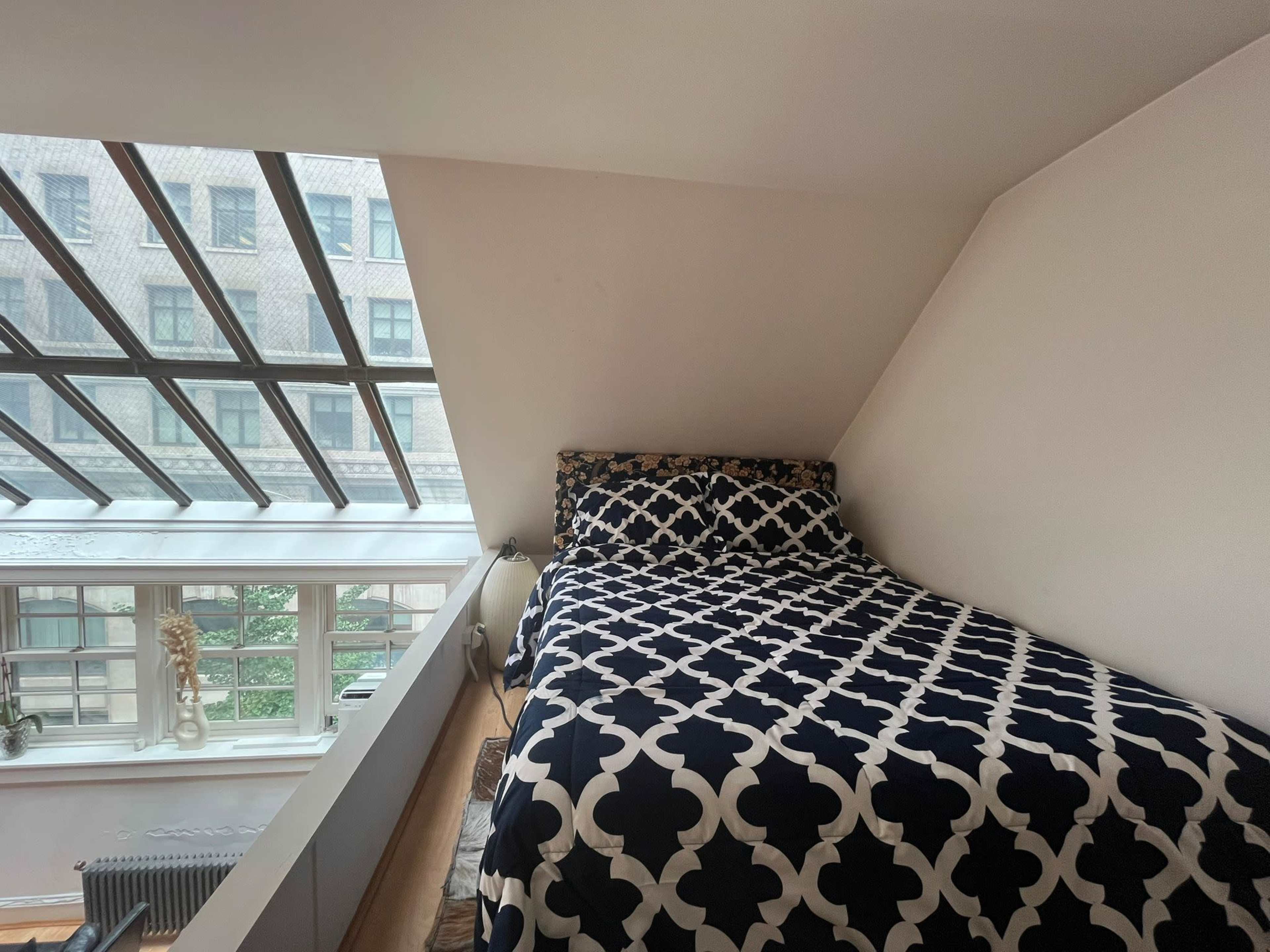 The scene features a bed with a patterned navy and white blanket, positioned under a sloped ceiling with large glass windows allowing natural light to enter the room.