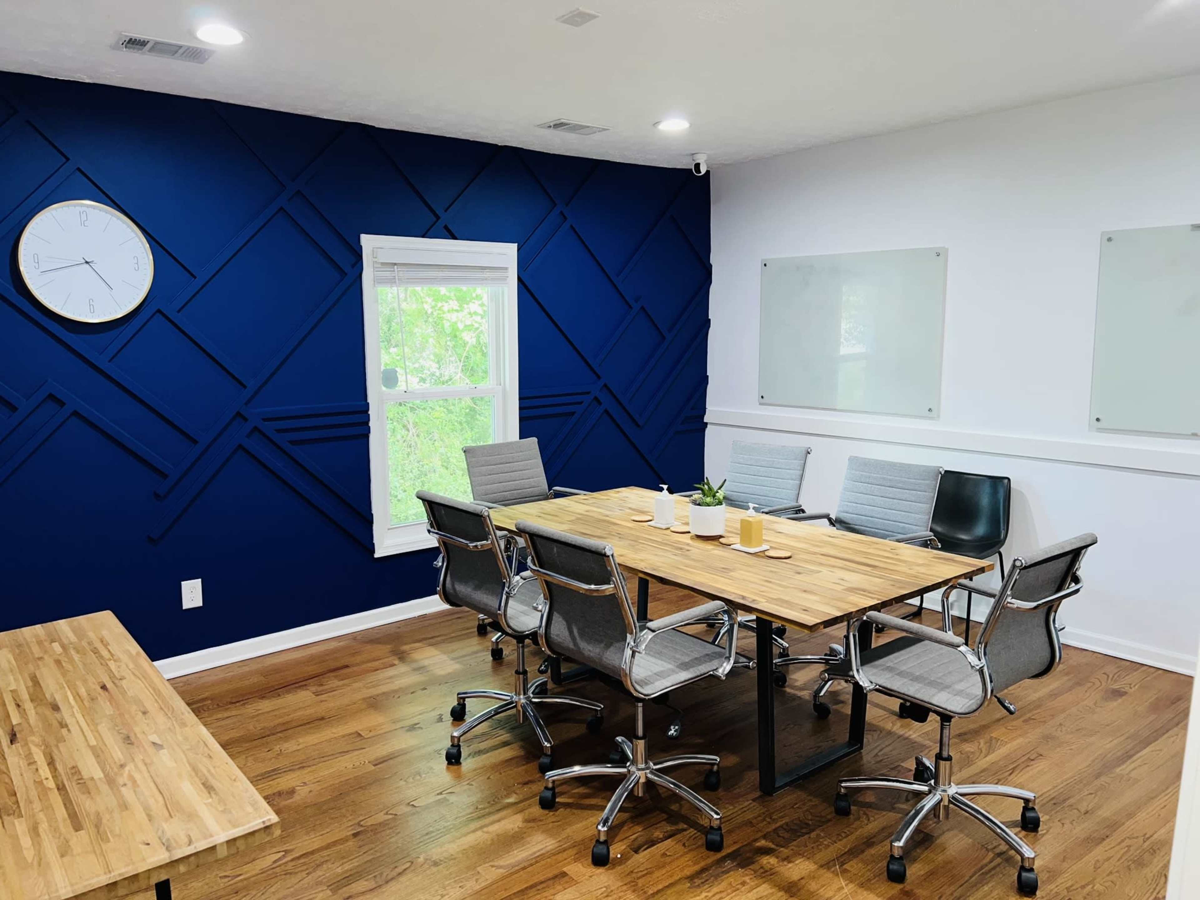 A modern conference room features a wooden table surrounded by ergonomic chairs, with a large clock on the wall and a blue accent wall behind it.