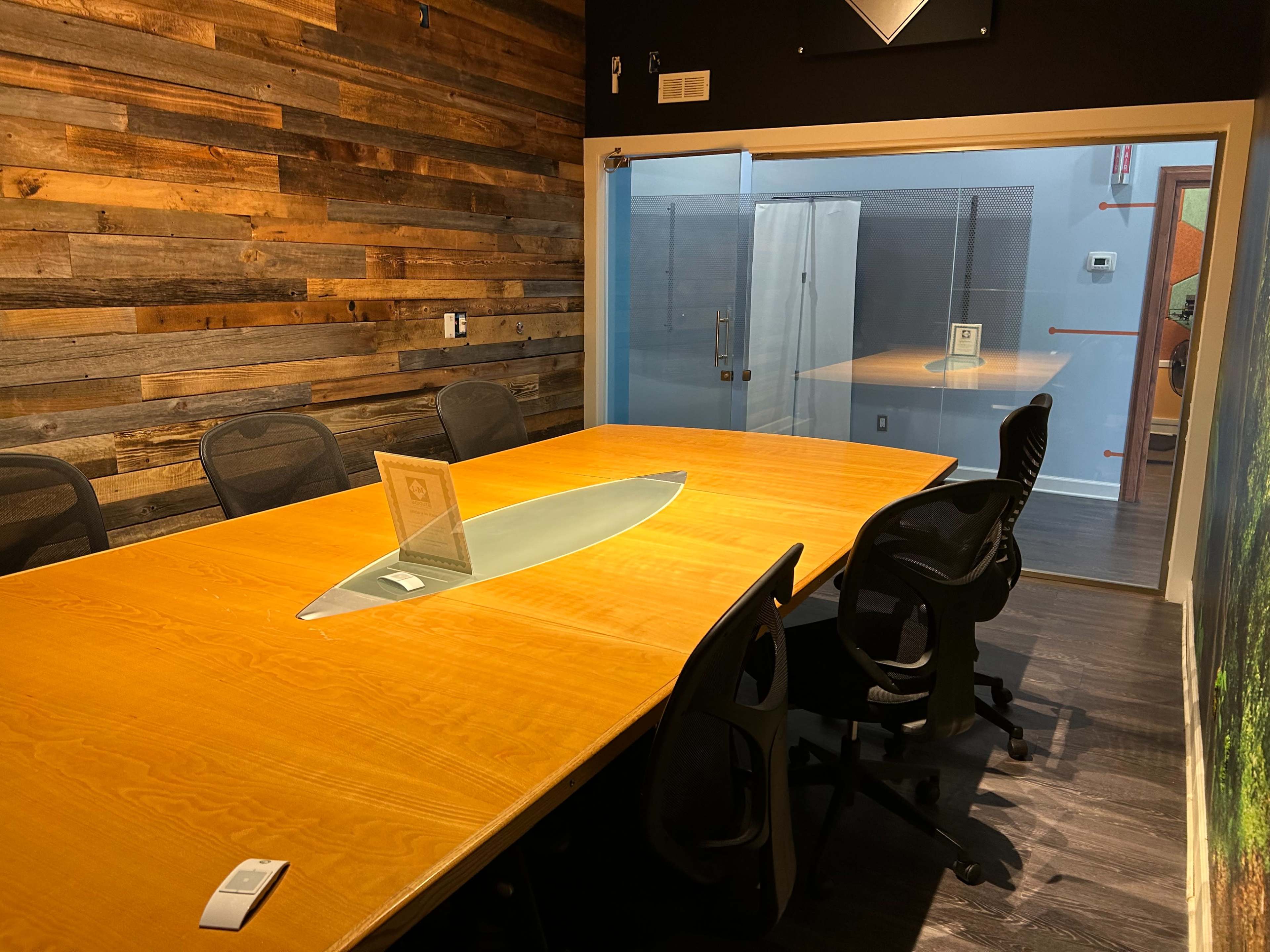 A modern conference room features a large wooden table with an oval glass centerpiece and black chairs arranged around it, adjacent to a glass-walled area.