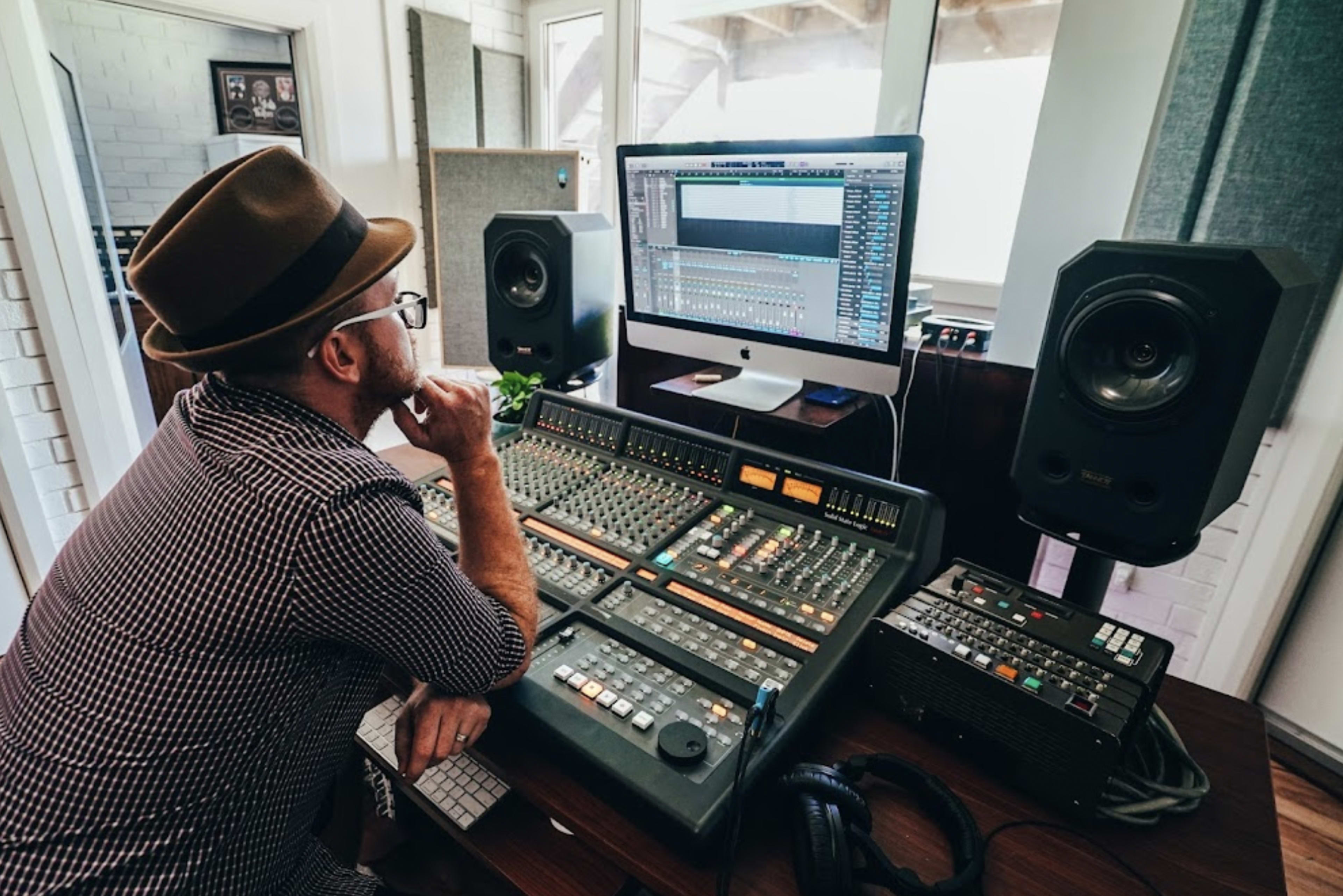 A man in a hat sits at a sound mixing console, focused on a computer screen displaying audio software in a professional recording studio.