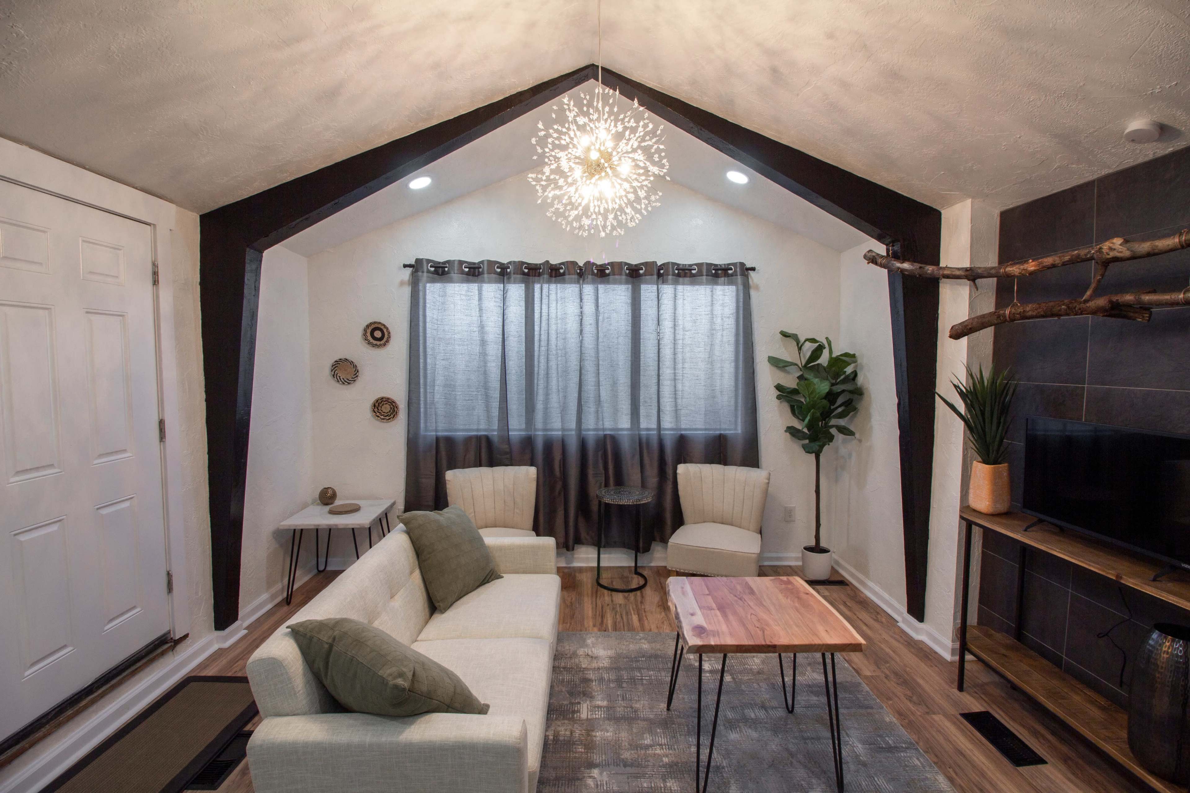 A modern living room features a sofa, two armchairs, a wooden coffee table, a decorative plant, and a wall-mounted TV, all illuminated by a distinctive pendant light and natural light from the window.