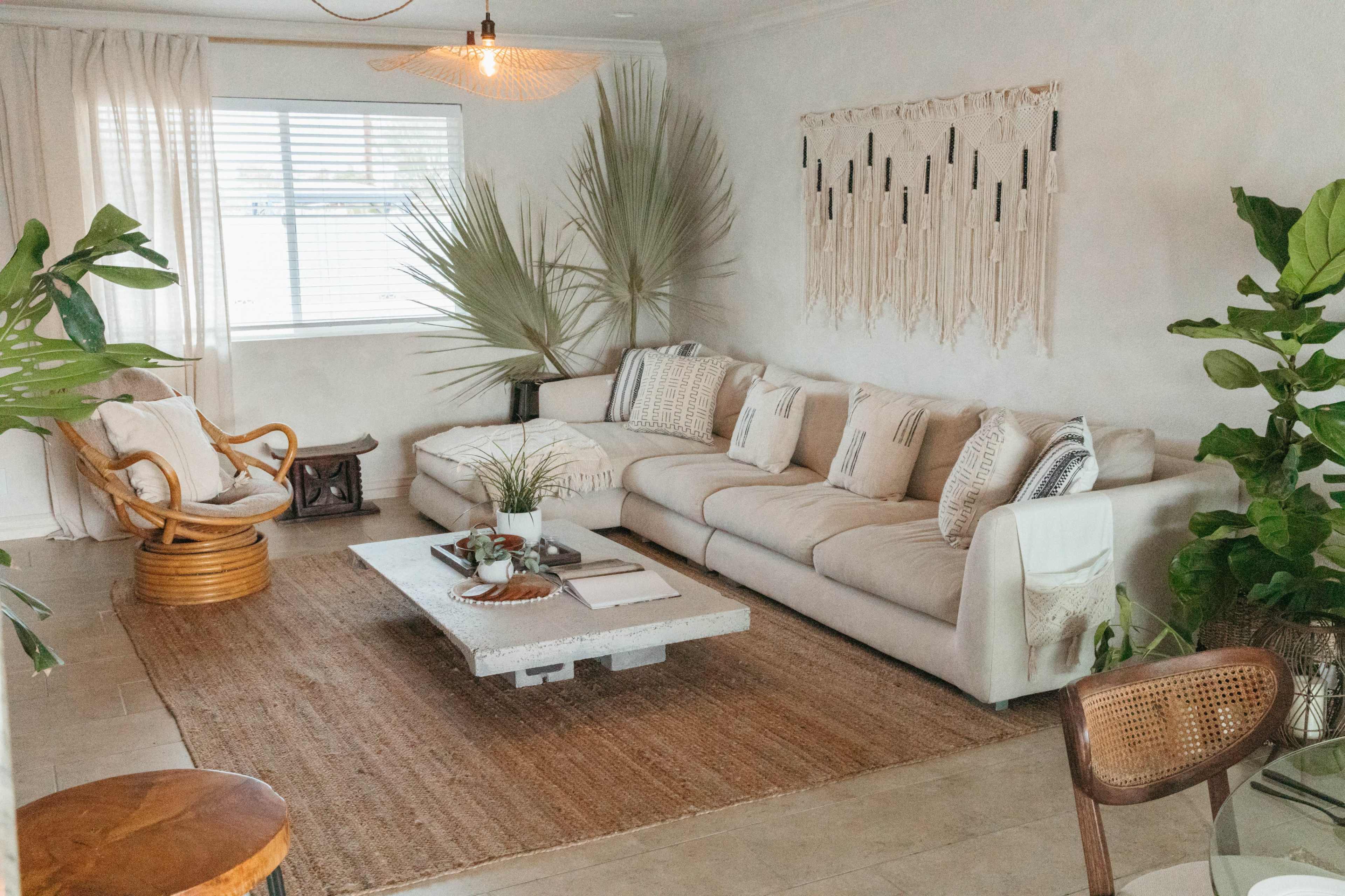 A cozy living room featuring a large light-colored sofa, a low coffee table, and various potted plants, with a woven rug on the floor and natural light coming through a window.