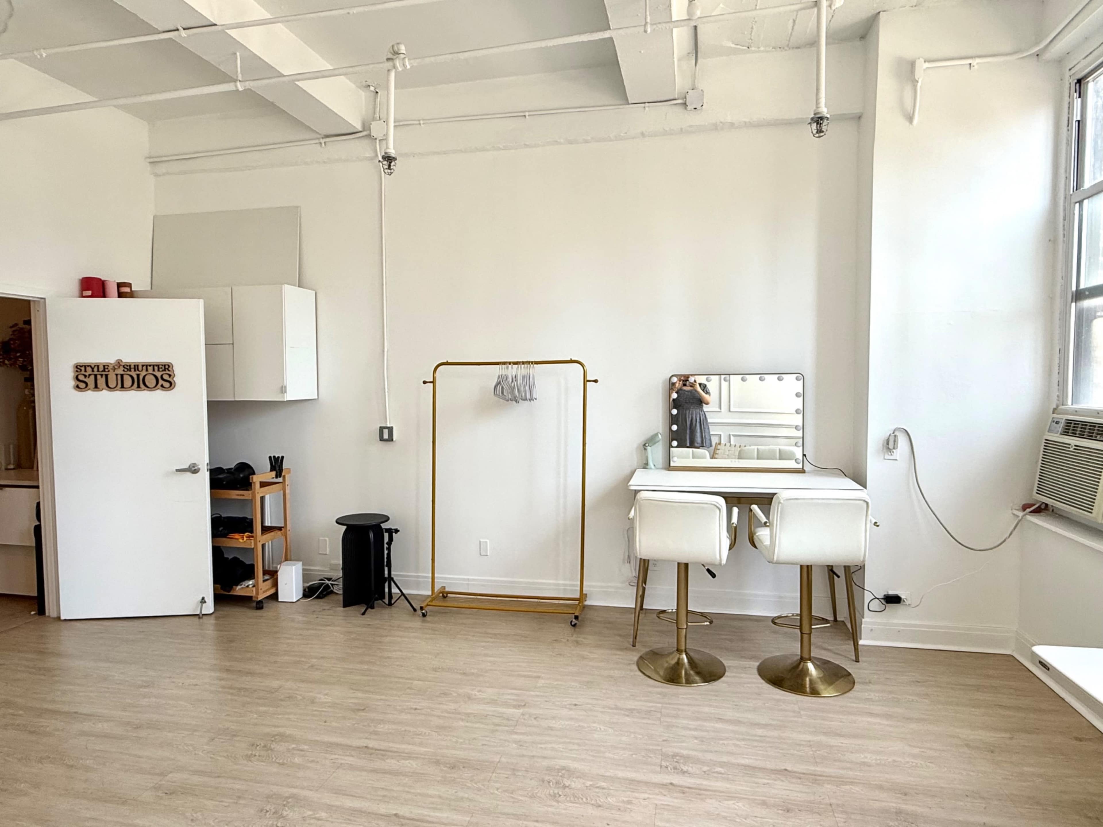 The image shows a bright, minimalist room with a clothing rack, a mirrored vanity with two stools, and an air conditioning unit near the window.