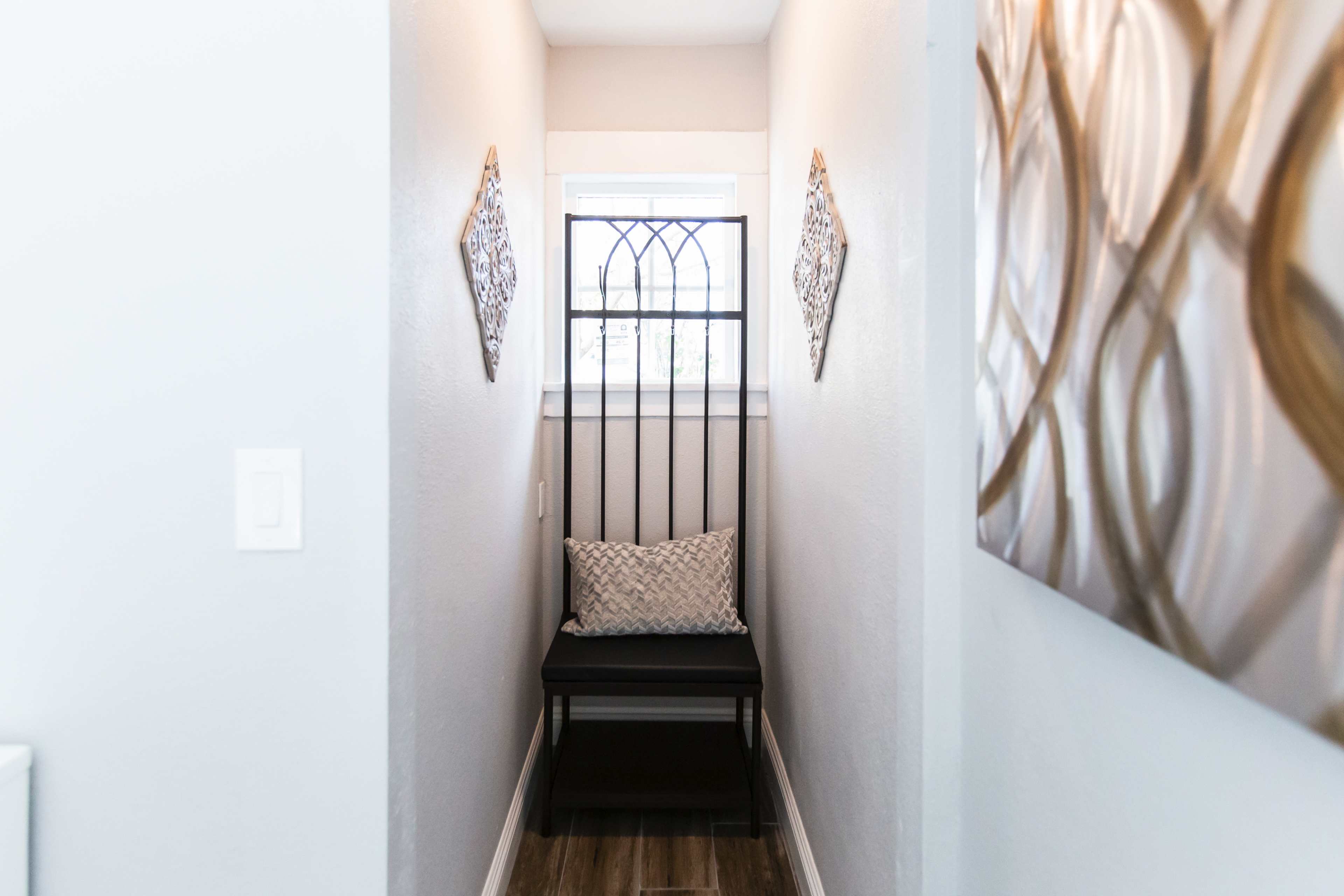 The image shows a narrow hallway featuring a bench against the wall, with decorative wall art on either side and a window at the far end.