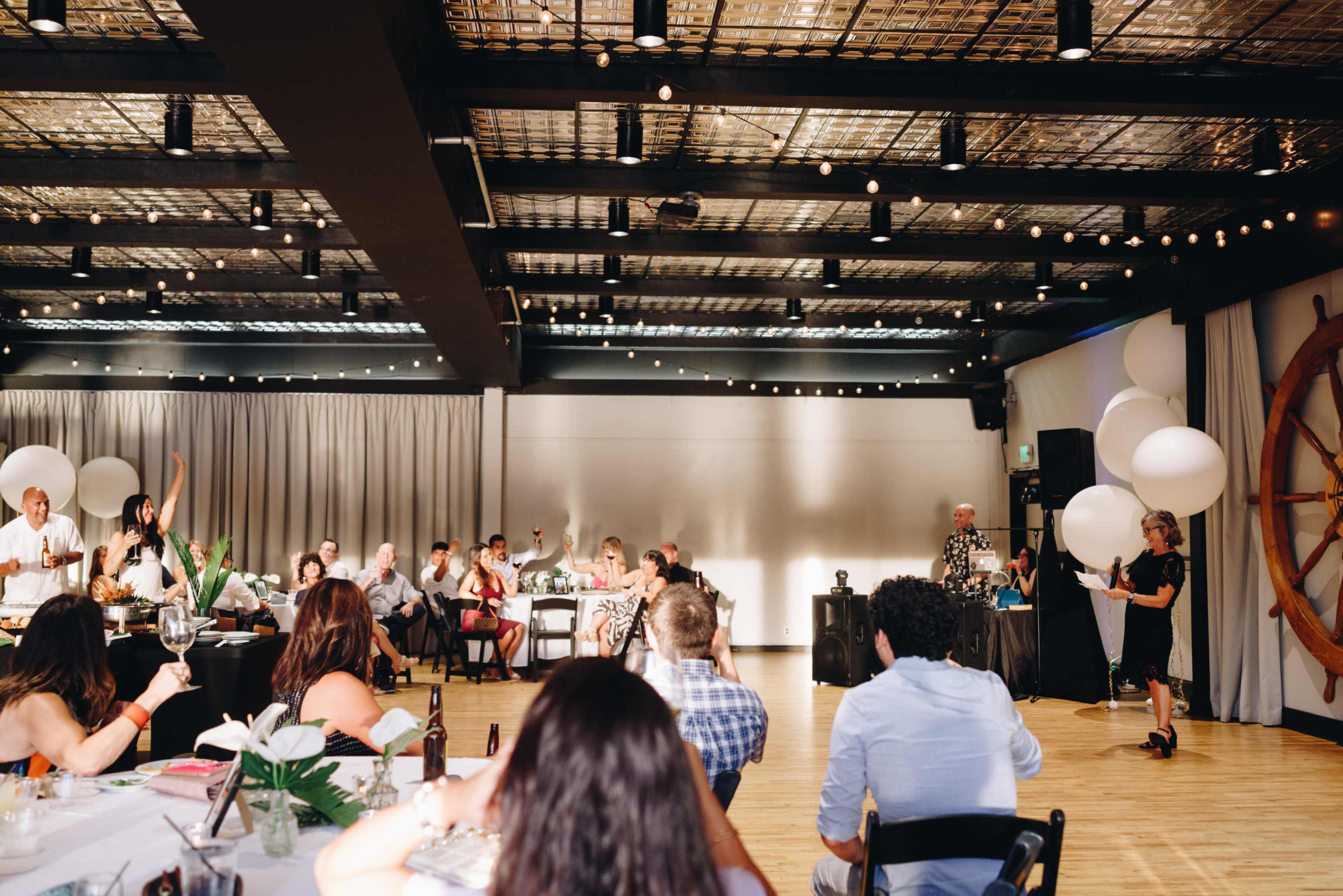 A large group of people is gathered in a well-lit indoor space decorated with balloons and plants, while some attendees listen to a speaker at the front.