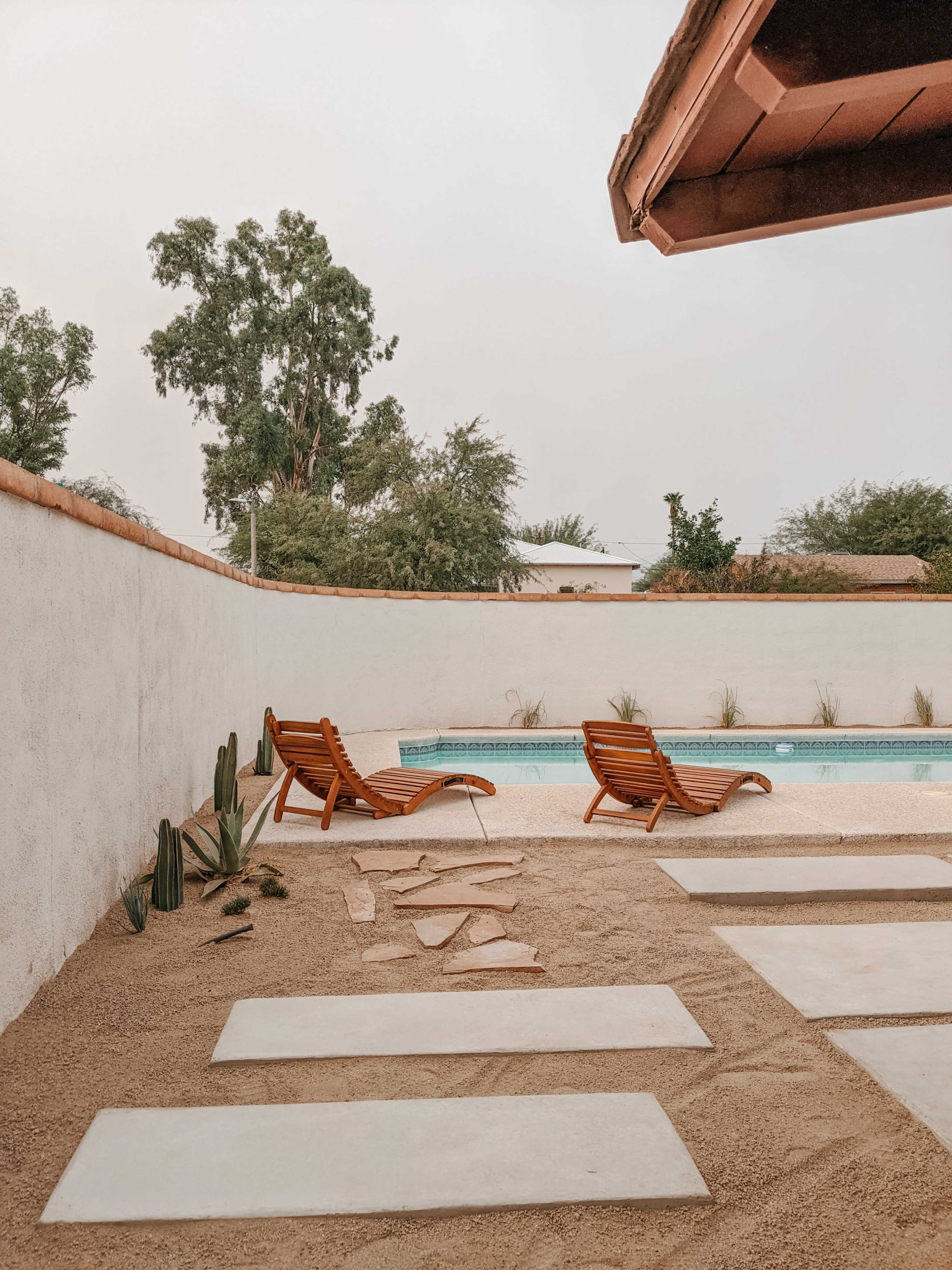 A pool area with two wooden lounge chairs, stone pavement, and cacti, surrounded by a white wall and greenery.