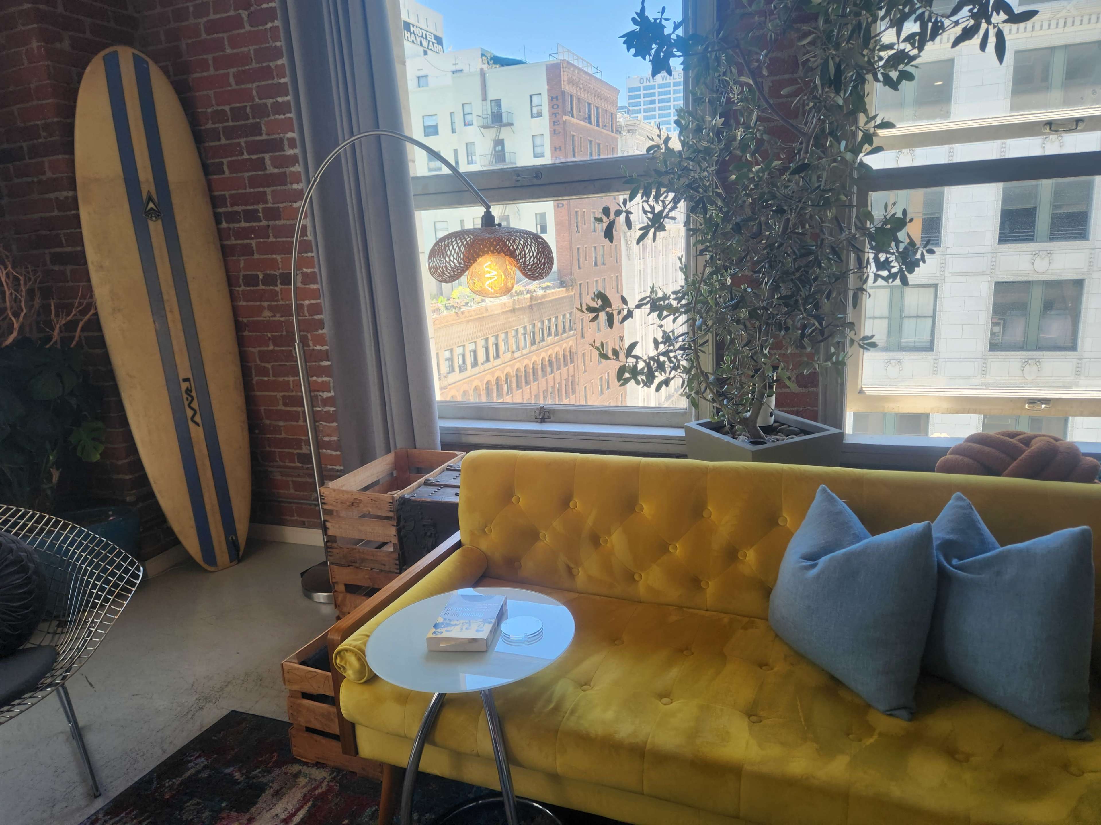 The scene features a yellow tufted sofa, a small side table with a book, a surfboard leaning against the wall, and large windows displaying a cityscape.