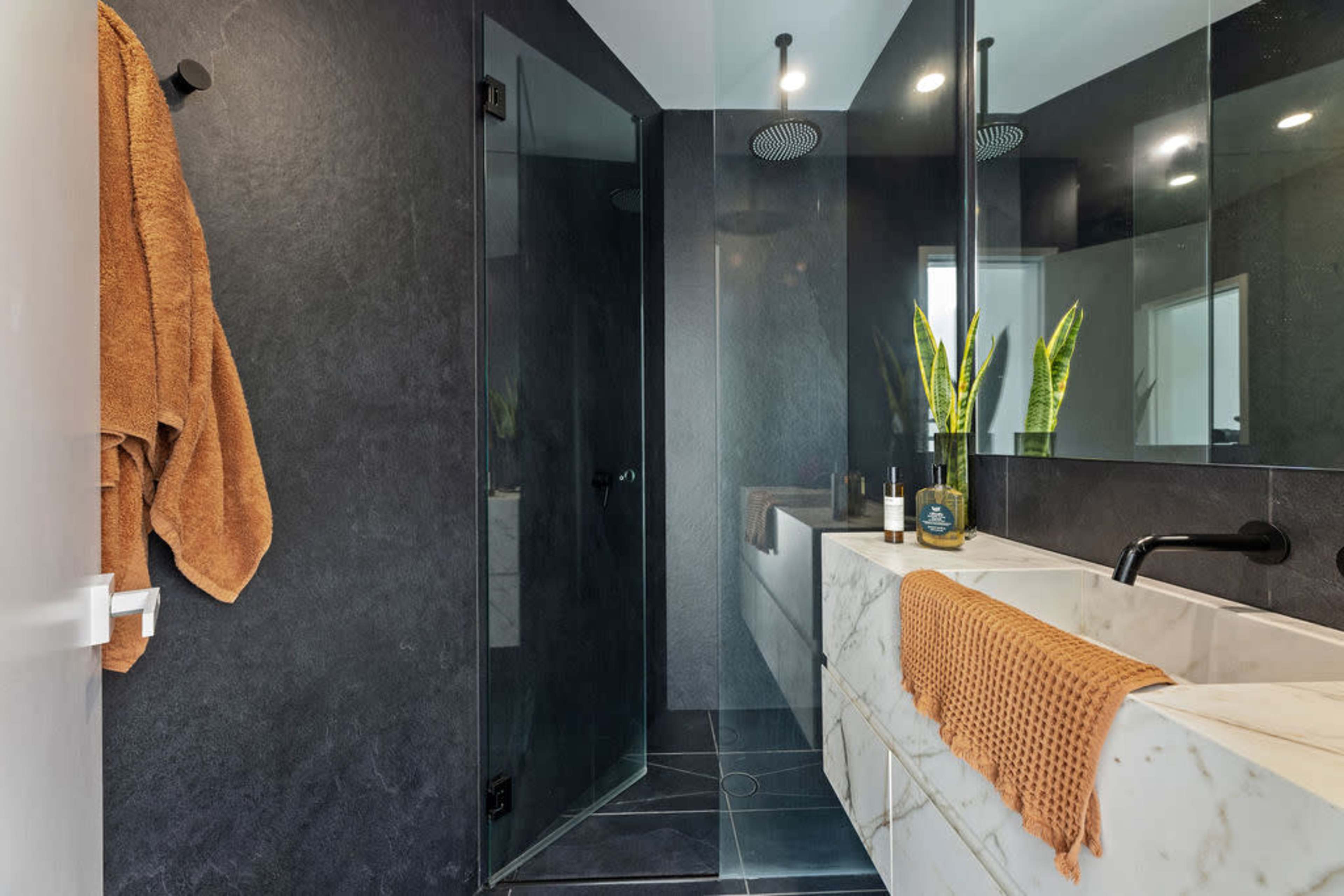 A modern bathroom features a walk-in shower with glass doors, a marble sink, and dark walls accented by green plants.