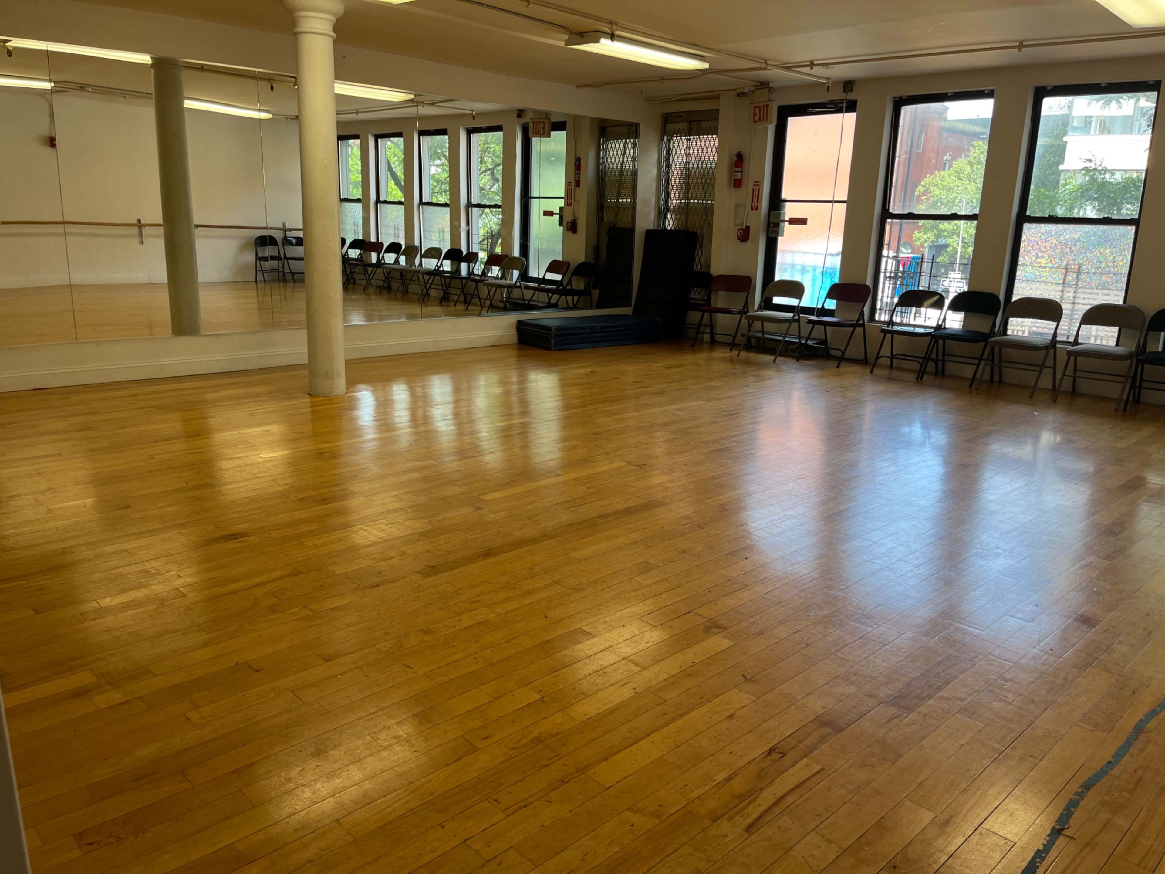 An empty dance studio with polished wooden floors, mirrors on one wall, and chairs lined up against another wall.