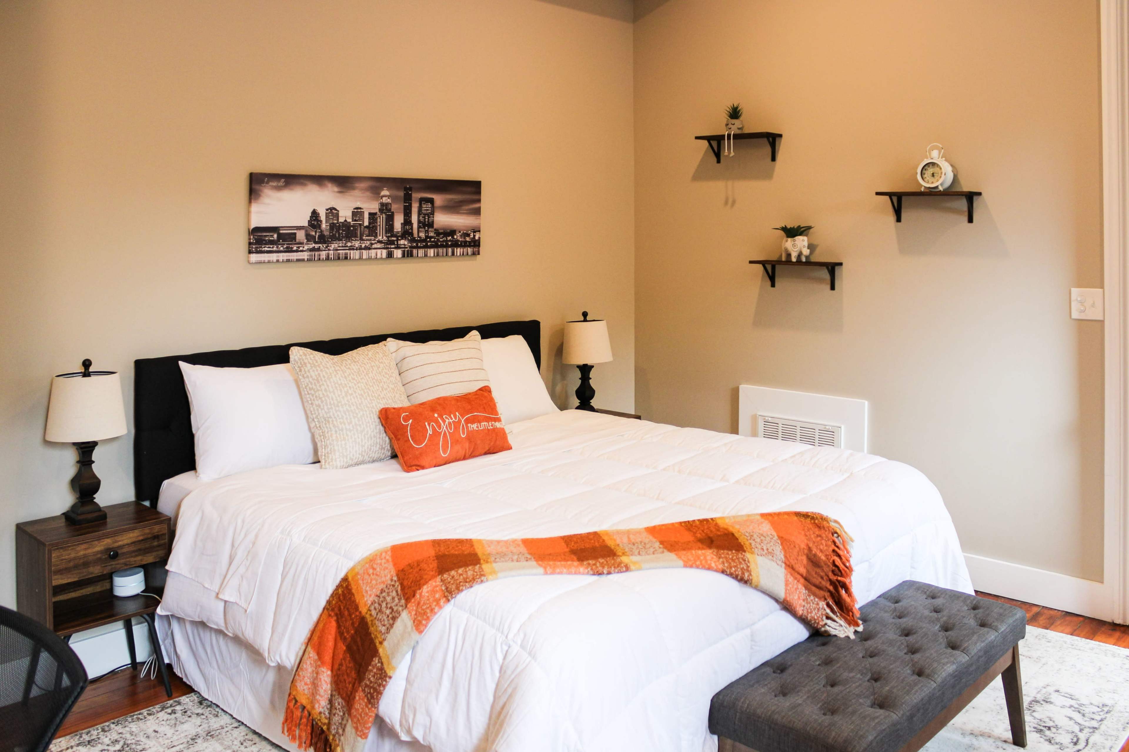 A cozy bedroom featuring a large bed with decorative pillows, an orange throw blanket, and minimalistic shelves on the wall.
