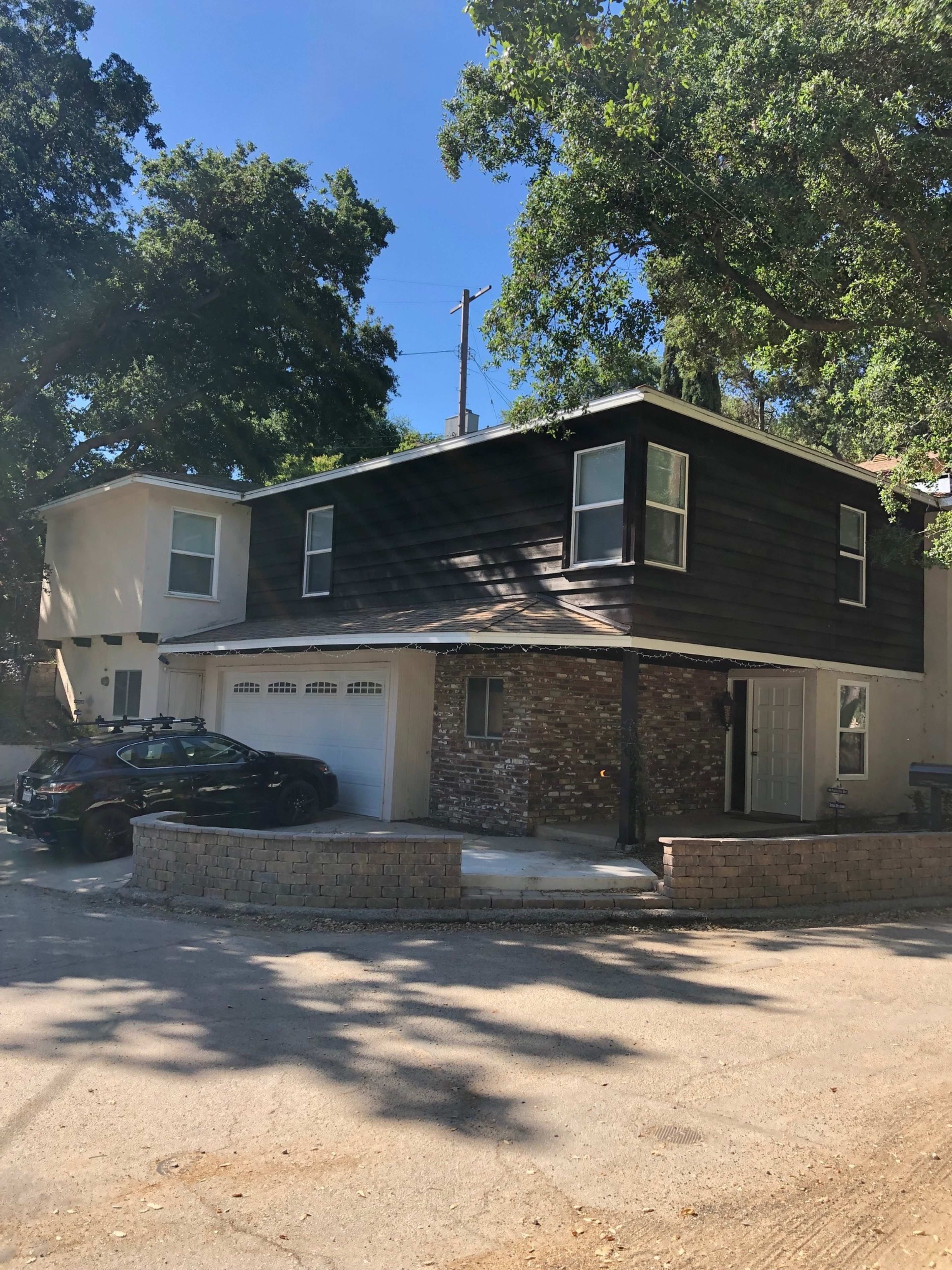 A two-story house with a mix of wooden and stone exterior is situated on a sloped driveway next to a smaller house, with a car parked in front.