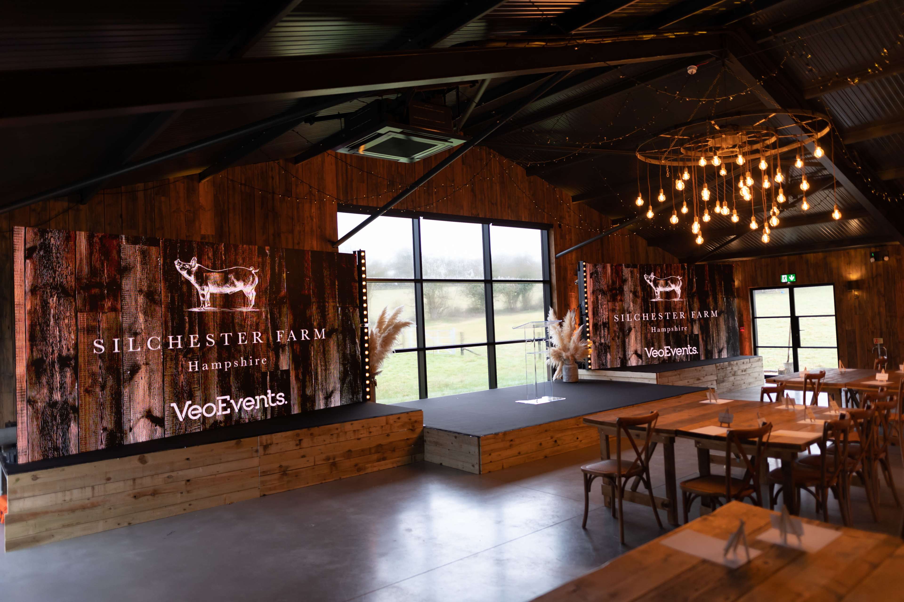 An event space at Silchester Farm in Hampshire, featuring a wooden stage and two large screens against a backdrop of rustic decor and hanging lights.