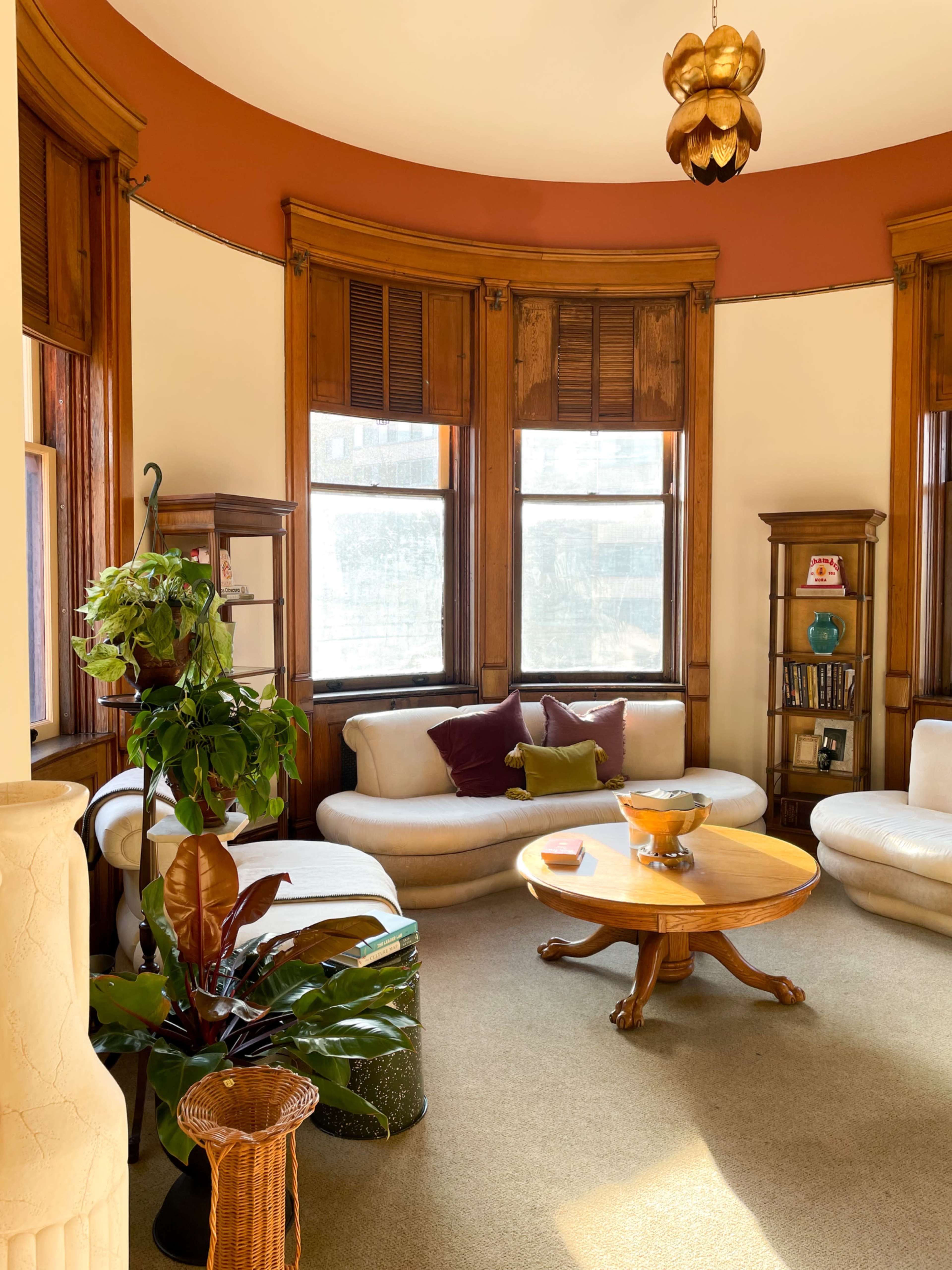 A semicircular room with large windows, featuring curved white sofas, a wooden coffee table, and potted plants.