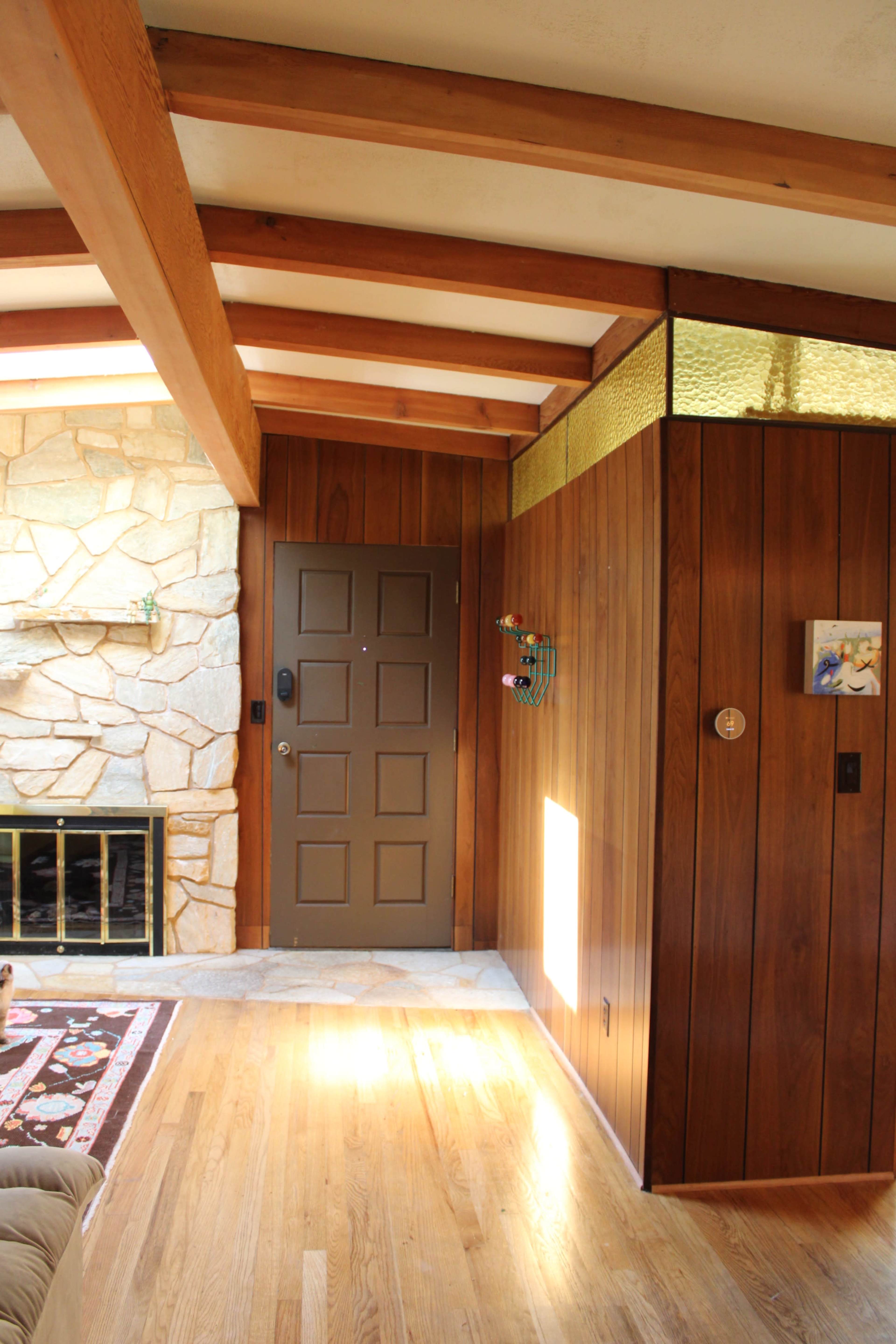 The image shows an interior hallway of a home with wooden walls, a stone fireplace to the left, and a dark brown door at the end of the hallway.