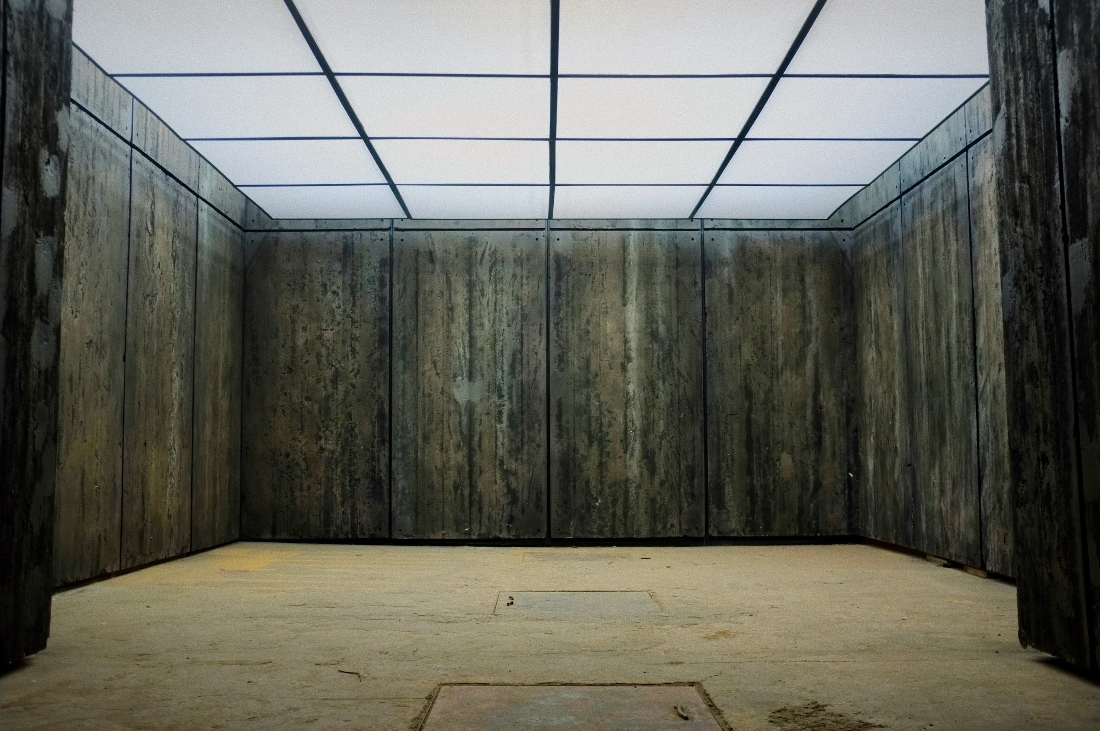 The image depicts an empty room with bare concrete walls and a grid-patterned ceiling.