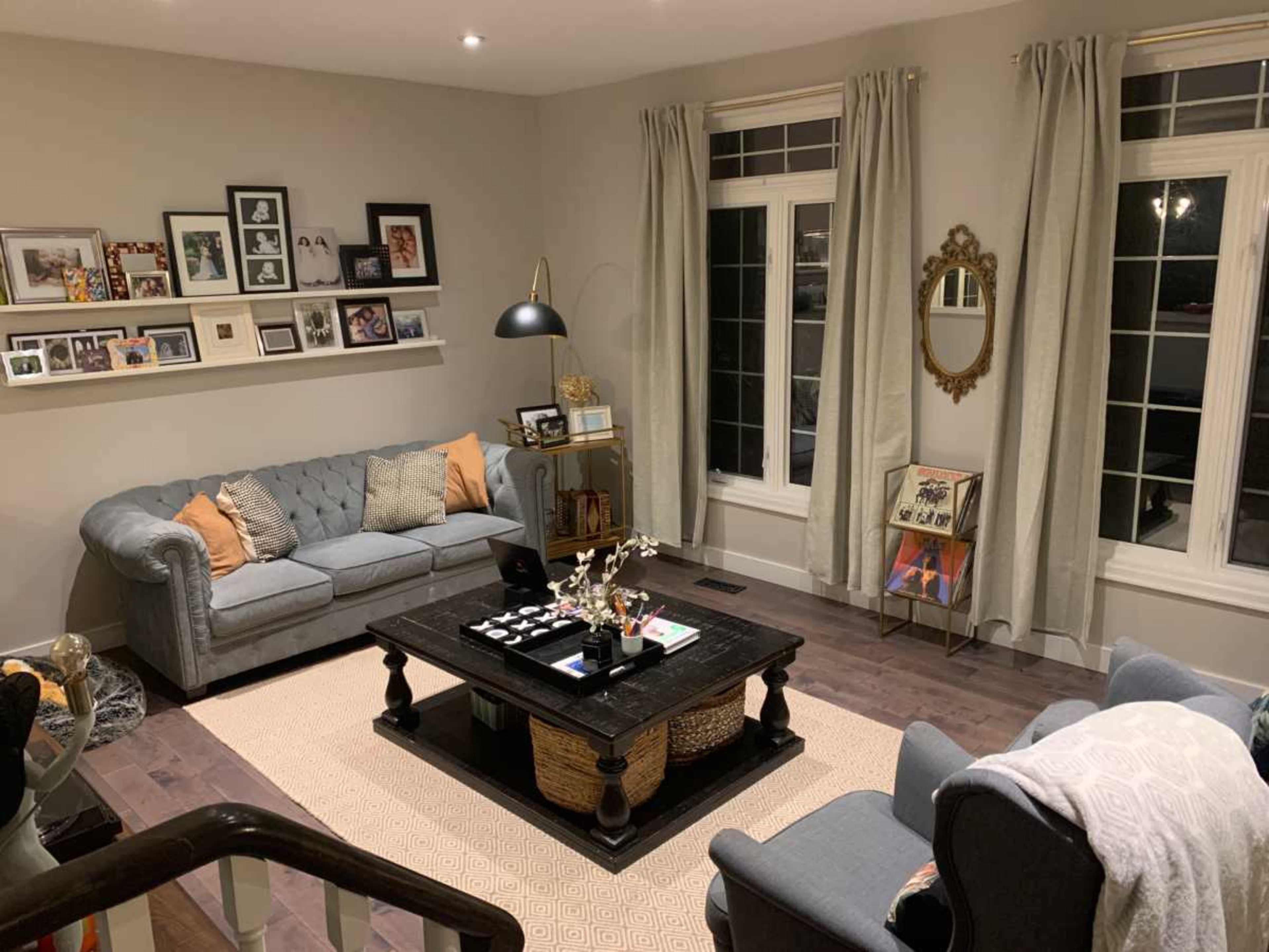 The image shows a cozy living room with a gray sofa, a coffee table, decorative items, and framed photos on the walls, illuminated by soft lighting through large windows.