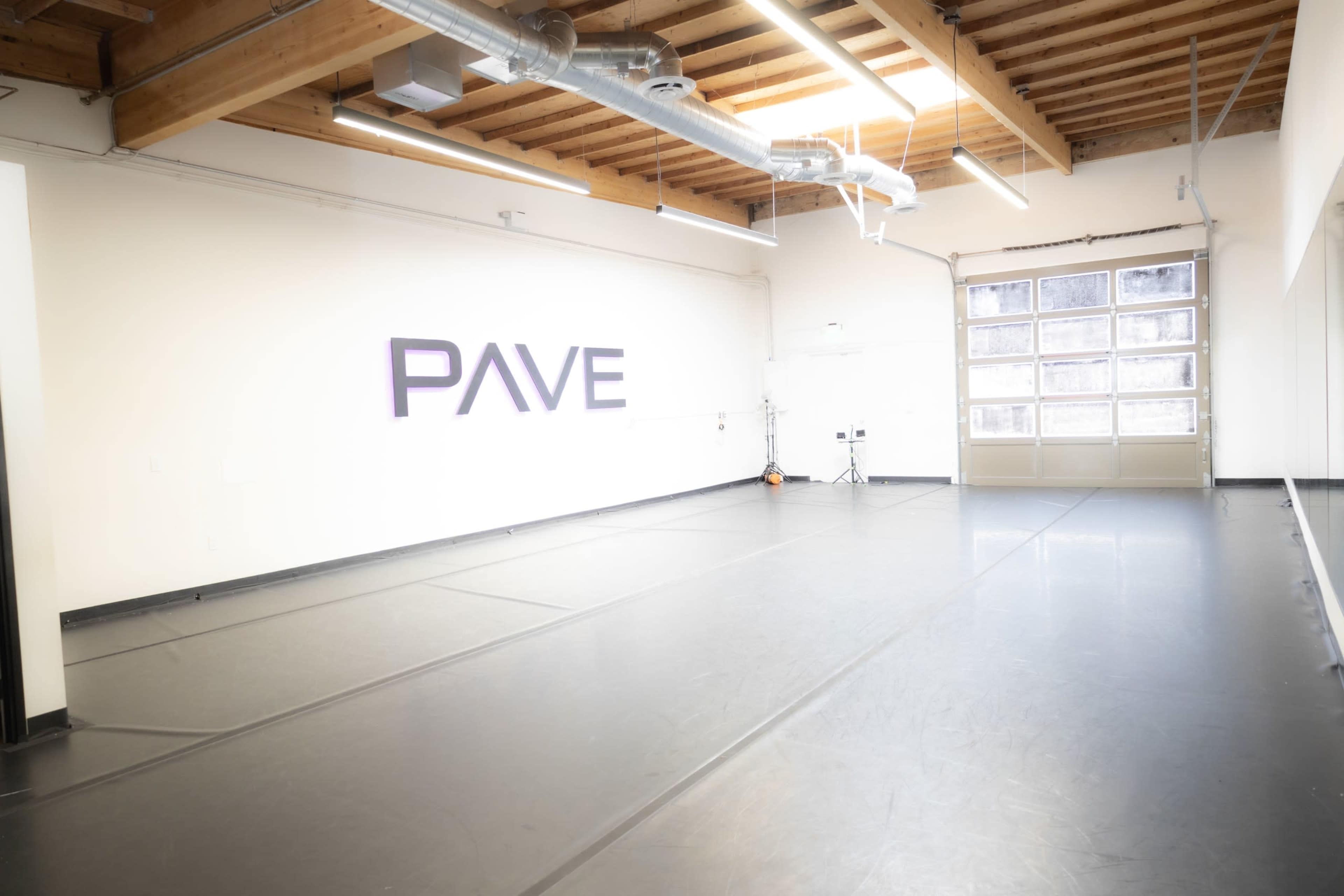 The image shows a spacious dance studio with a smooth black floor, high ceilings, and a large window allowing natural light.