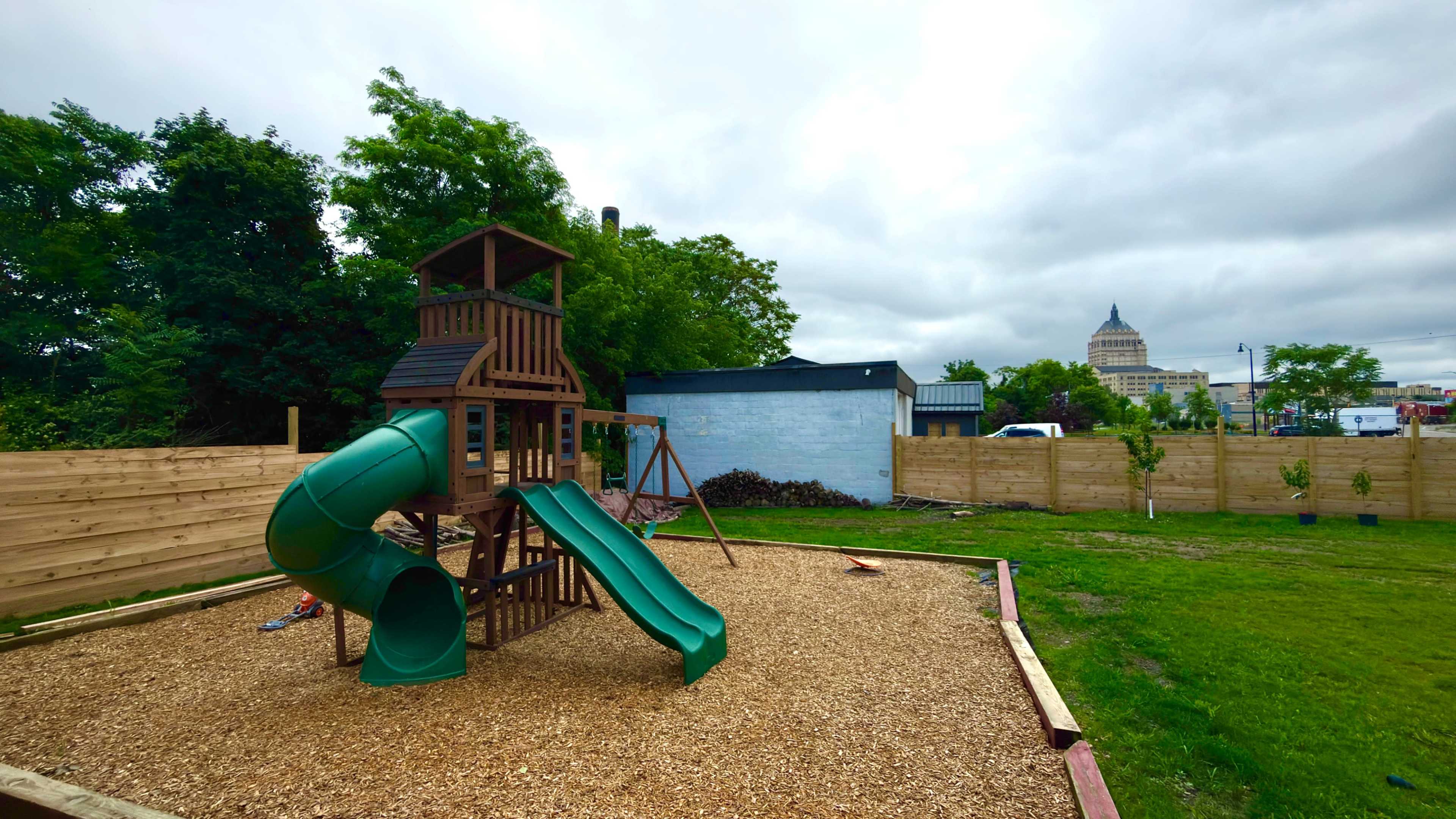 The image shows a wooden playground structure with slides and a climbing frame situated on a bed of wood chips, surrounded by a grassy area and fences, with a building and a tower visible in the background under a cloudy sky.