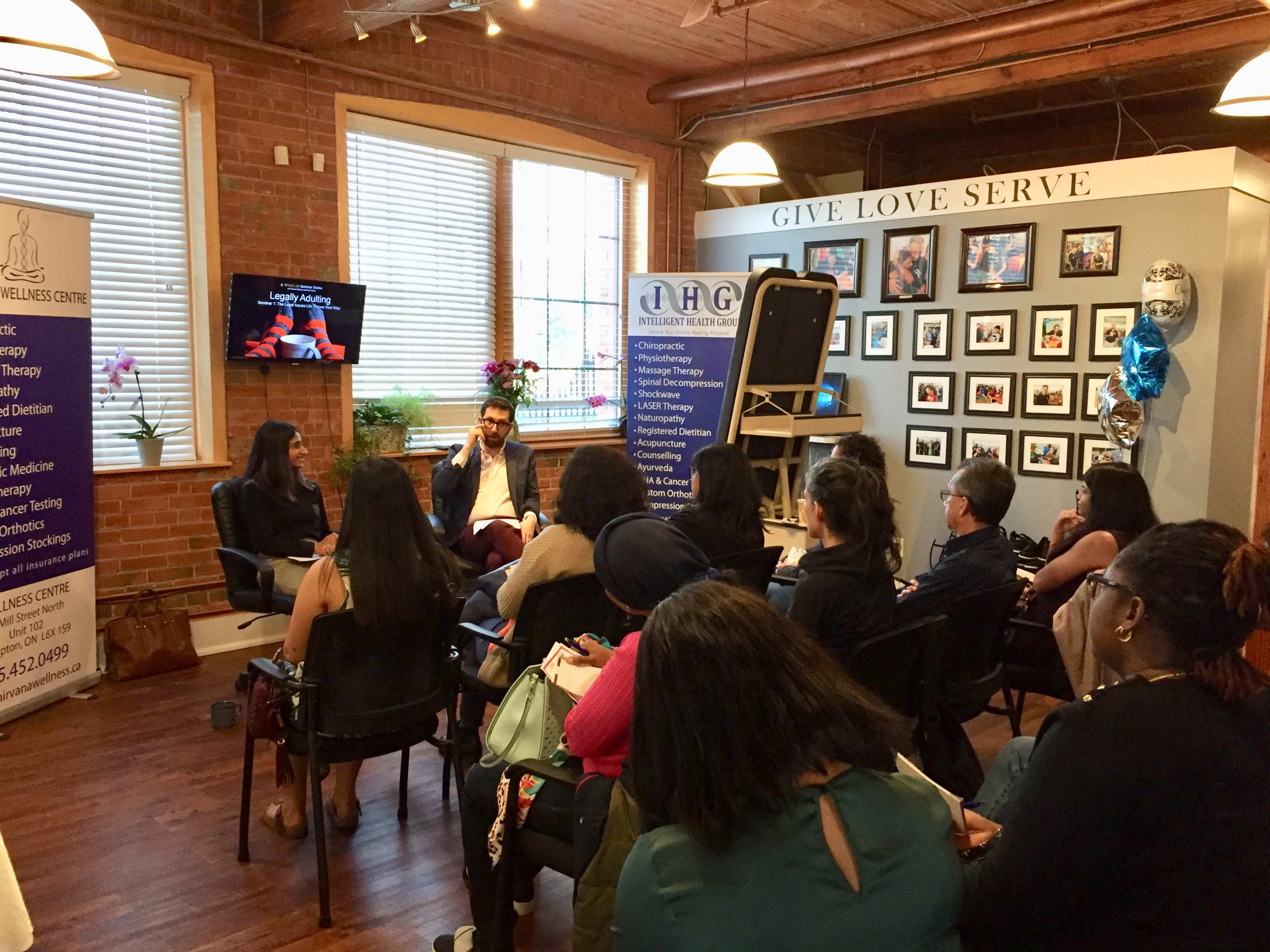 Two speakers engage with an audience in a room adorned with photographs and decor, while participants listen attentively from their seats.
