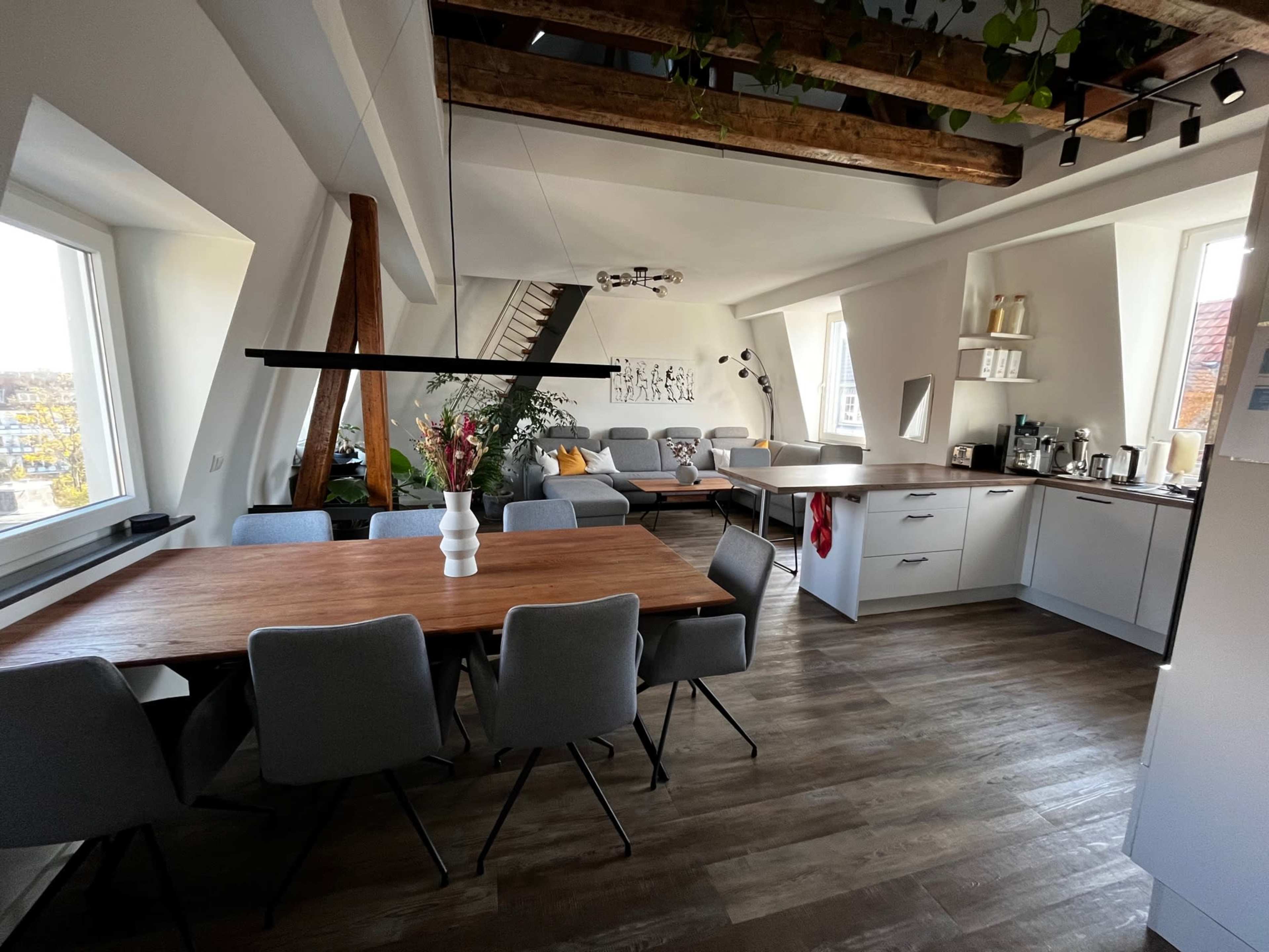 A modern kitchen and dining area featuring a wooden table with gray chairs, white cabinetry, and large windows providing natural light.