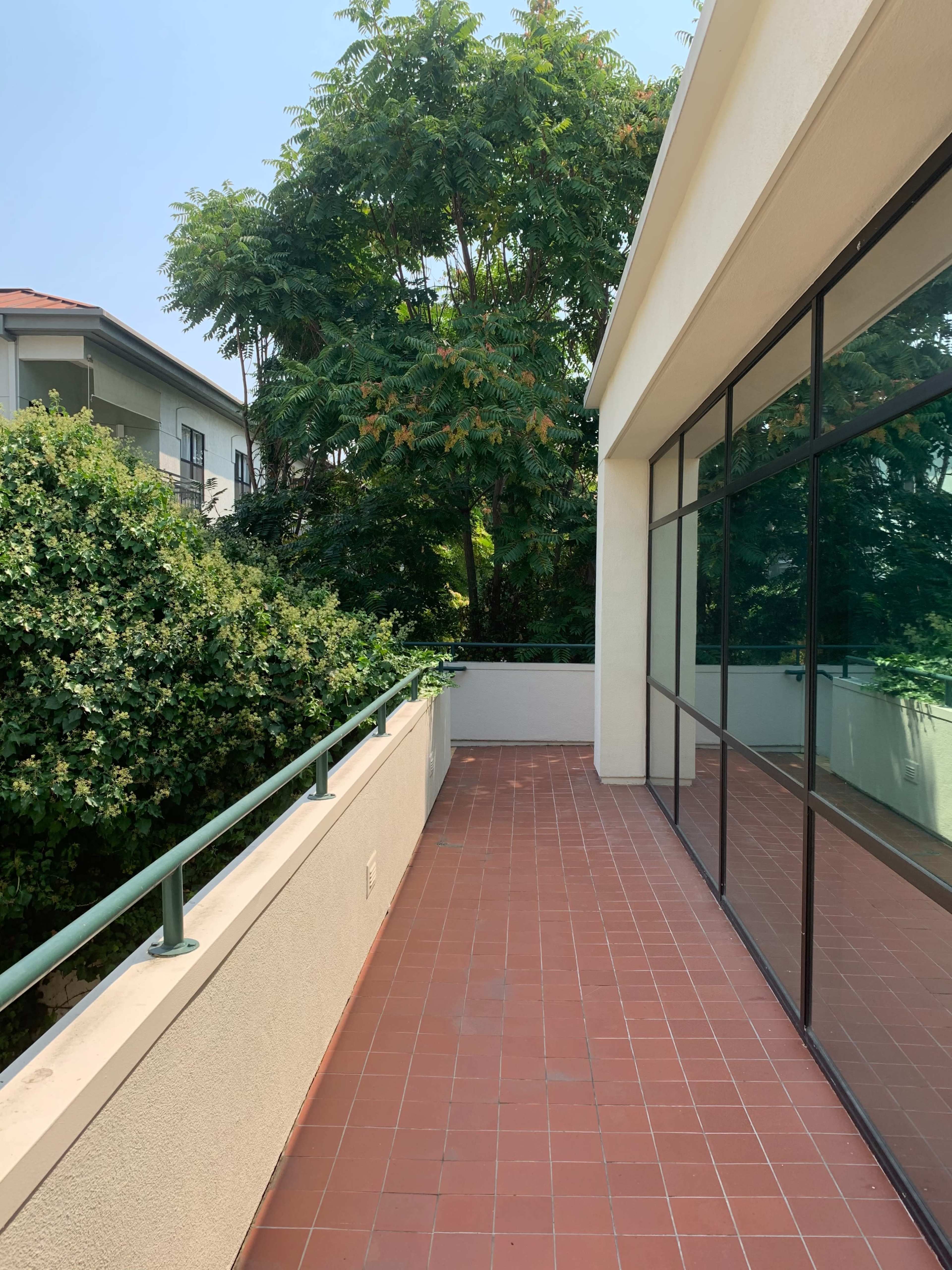 The image shows a tiled outdoor balcony with greenery and a large glass wall reflecting the surroundings.