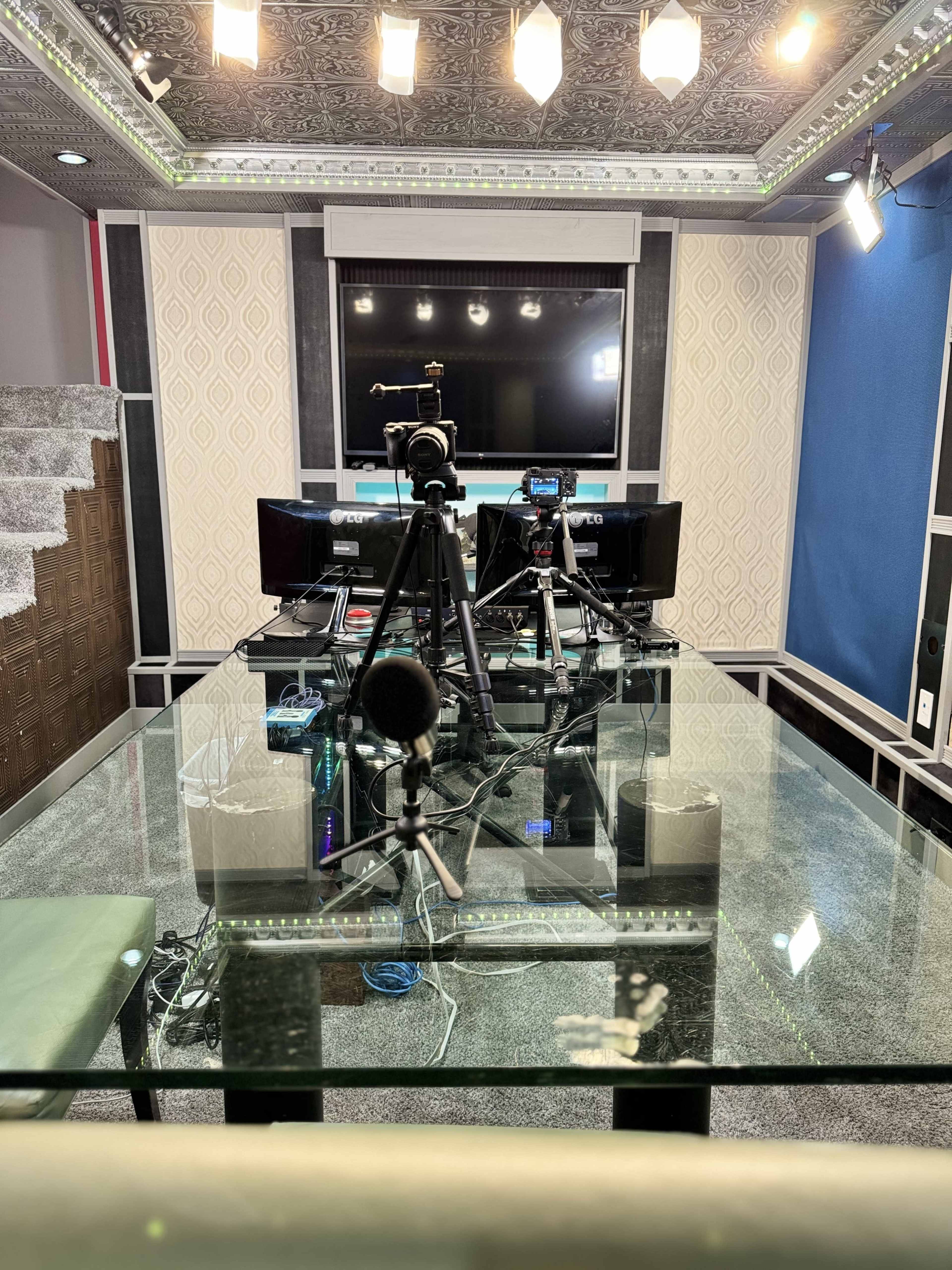 A modern studio setup featuring a glass table with multiple cameras and microphones positioned in front of two computer monitors.